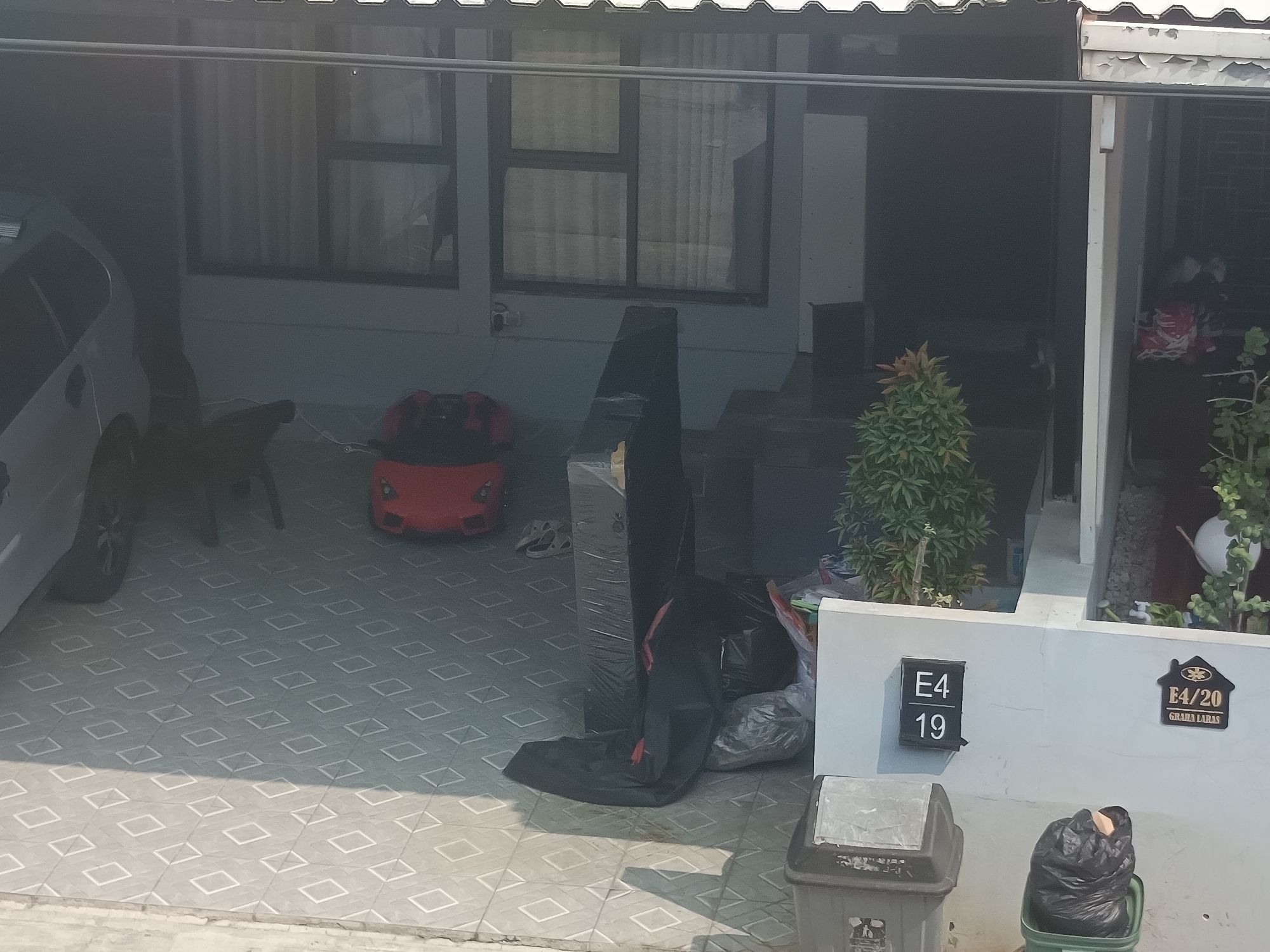 The image depicts the exterior of a residential building in Sukaraja, Indonesia, likely a carport or front patio area. The floor is tiled with a diamond pattern in a light gray color. To the left, a portion of a white car is visible, with its rear wheel on the tiled floor. In the center, a red toy car is parked on the tiled surface. Adjacent to the toy car, a black plastic bag is wrapped around a tall, rectangular object, possibly furniture or packaging. Further to the right, a small, conical green plant is situated in a planter. On the wall behind the plant, a black house number sign reads "E4 19". To the right of this sign, another sign, shaped like a house, displays "E4/20" and "GIRANA LAPAS" in gold lettering. In the foreground, two trash bins are visible: a grey one with a partially open lid on the left, and a dark green one on the right containing a black trash bag. Sunlight casts shadows across the tiled floor, suggesting it is daytime. The overall impression is of a residential entryway with various items stored outdoors.