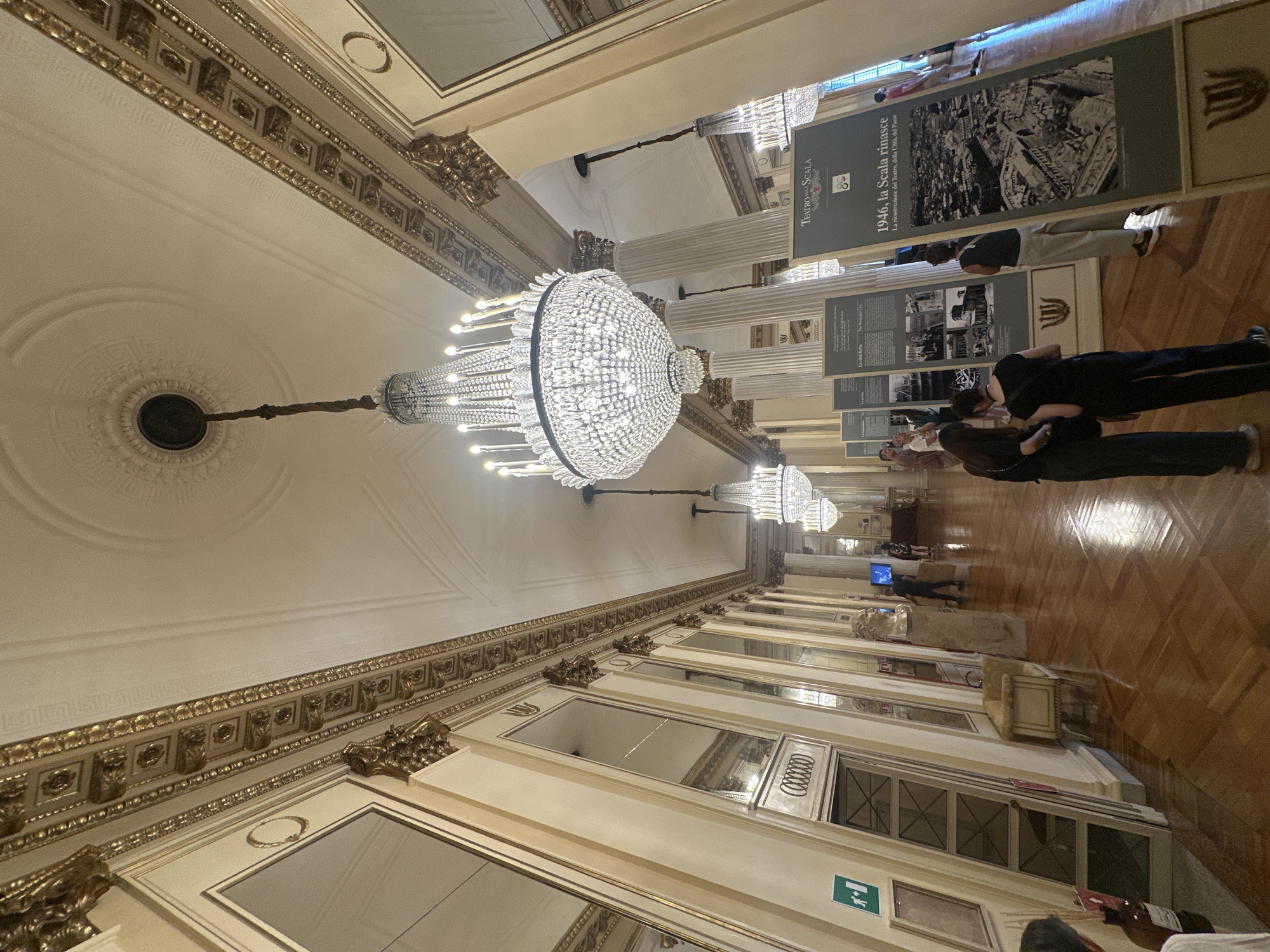The image shows a grand hallway with high ceilings and ornate gold detailing. Several large, multi-tiered crystal chandeliers hang from the ceiling, illuminating the space with a warm glow. The walls are lined with tall mirrors, reflecting the ornate architecture and the chandeliers. There are classical columns along the hallway, and the floor is made of polished dark wood in a parquet pattern.
In the foreground, a group of people are looking at display boards. These boards appear to be informational exhibits, with text and images. One prominent display board in the center right has the title "1946, la Scala rinasce" and a black and white aerial photograph of a city.
Further down the hallway, more people are visible, some walking and others looking at exhibits. An exit sign with a green running figure is visible on the left wall. The overall impression is one of a well-maintained, elegant interior, likely a museum, gallery, or historic building. The location is identified as the Teatro alla Scala in Milan, Italy.