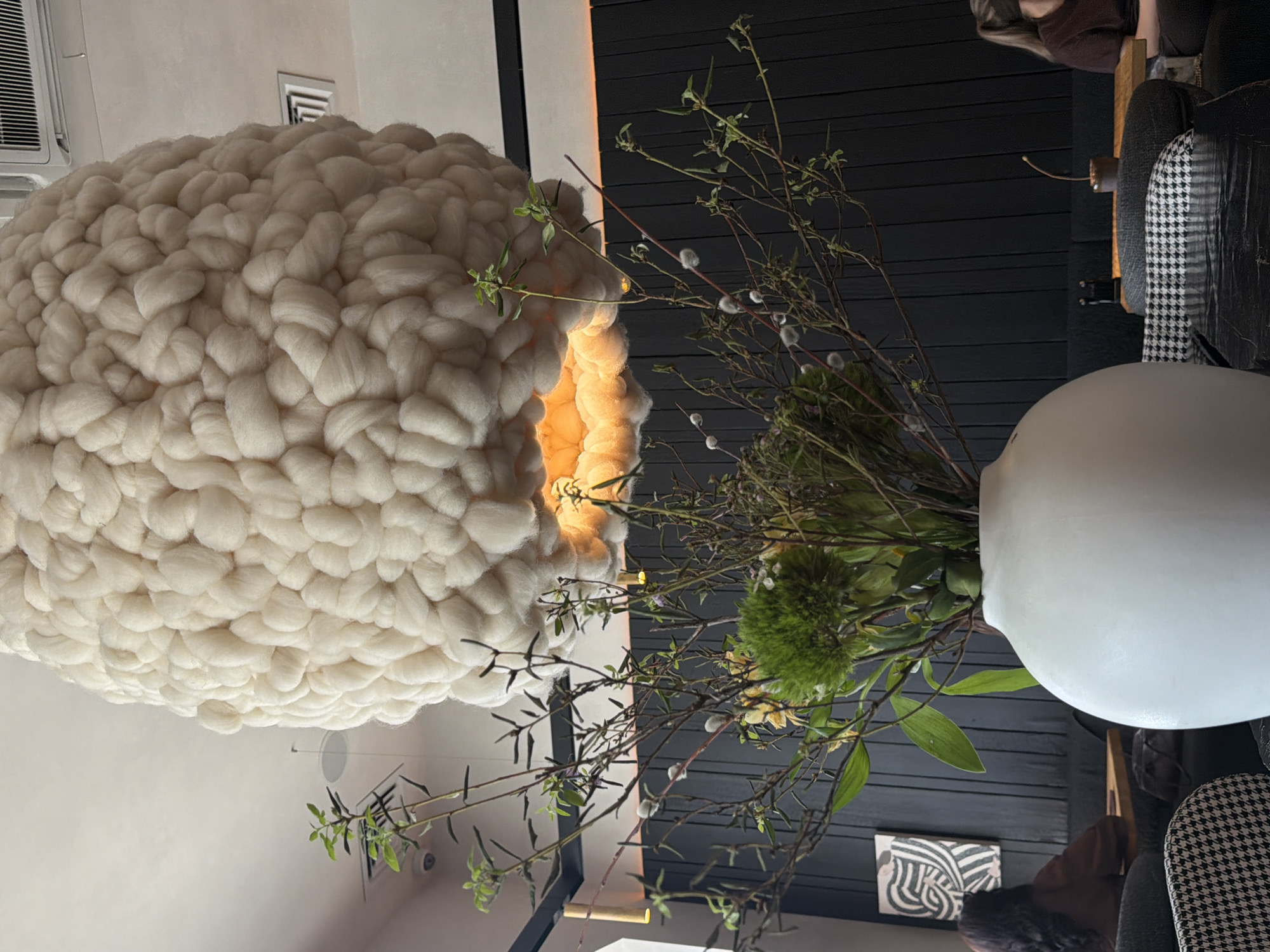 The scene is inside a modern-designed room featuring a large, cloud-like chandelier made of intertwined white woolen material, hanging from the ceiling. A white spherical vase filled with green foliage and slender branches, some with pussy willows, sits on a table. Behind the vase, there's a dark, horizontally paneled wall. In the background, a houndstooth-patterned chair, and a person in brown attire are visible, possibly seated. A framed piece of abstract art is on the wall. The image provides no information to determine whether the scene is located in Krasnoyarsk, Russia.