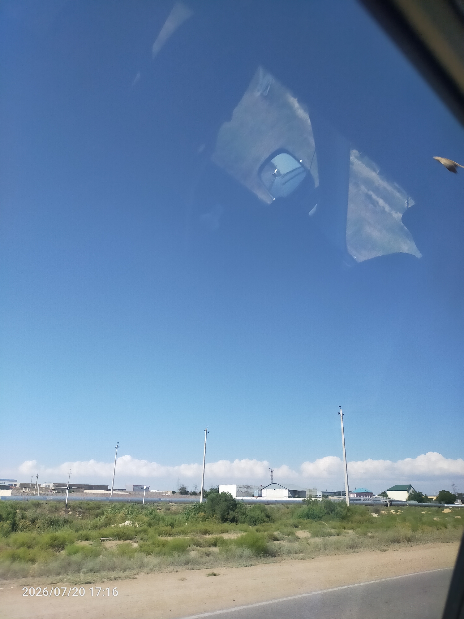 The image shows a bright, clear blue sky with reflections on a surface that appears to be a window. In the upper portion of the image, there are reflections of what seem to be parts of a vehicle interior, including a rearview mirror. A bird is visible flying in the upper right quadrant of the sky. The lower portion of the image displays a landscape with sparse vegetation, including dry grasses and bushes, with a dirt road in the foreground. In the background, there are utility poles, buildings with light-colored roofs, and a fence. A date and time stamp, "2026/07/20 17:16", is visible in the lower left corner of the image.