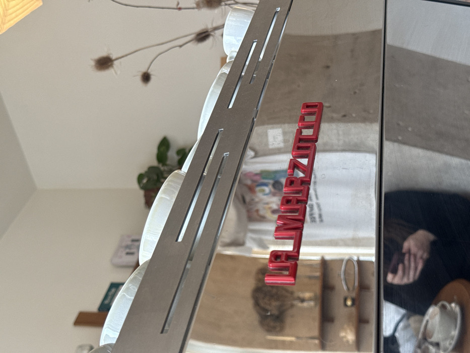 This image captures a close-up view of the reflective stainless steel facade of a La Marzocco espresso machine within a cafe in Paris, France. A red, three-dimensional "LA MARZOCCO" logo is prominently affixed to the machine's surface. Several white ceramic coffee cups are arranged on the machine's upper warming tray. The polished surface reflects the cafe's interior: a person wearing a light-colored t-shirt with a colorful graphic design is visible in reflection, standing in front of a light-colored wall. To the reflected person's left, a wooden shelf with decorative items and a green potted plant are visible. To the right, dried thistles in a vase and a woven light fixture are reflected. In the lower portion of the reflection, a person dressed in dark clothing, holding a smartphone in their right hand, is observed taking the photograph. Adjacent to this reflection, a portion of a wooden table is visible, upon which rests a white coffee cup containing a beverage and a matching saucer.