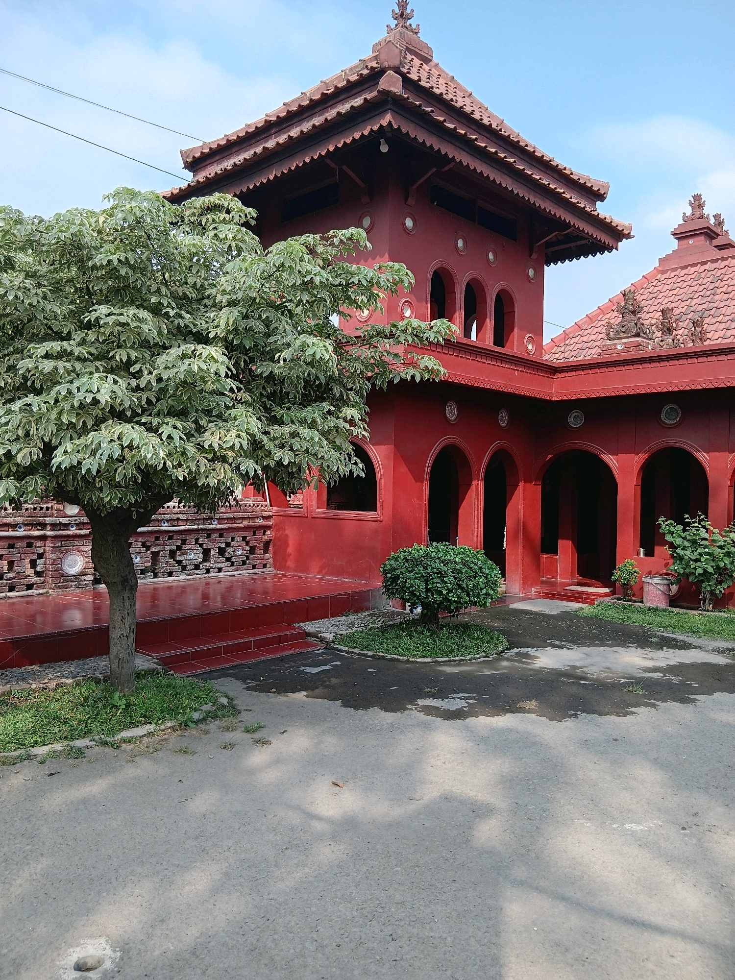 The image shows the exterior of the Sunyaragi Building in Cirebon, Indonesia, under a clear blue sky. The building is predominantly painted a vibrant red and features traditional Javanese architectural elements, including tiered roofs with intricate trim and multiple arched doorways. A large tree with variegated green and white leaves stands to the left of the building, partially obscuring a red tiled walkway and a patterned brick wall. In the foreground, a gray asphalt path is visible, with patches of grass and scattered leaves. The scene is static, with no people or animals present. The overall impression is one of a historical and culturally significant site.
