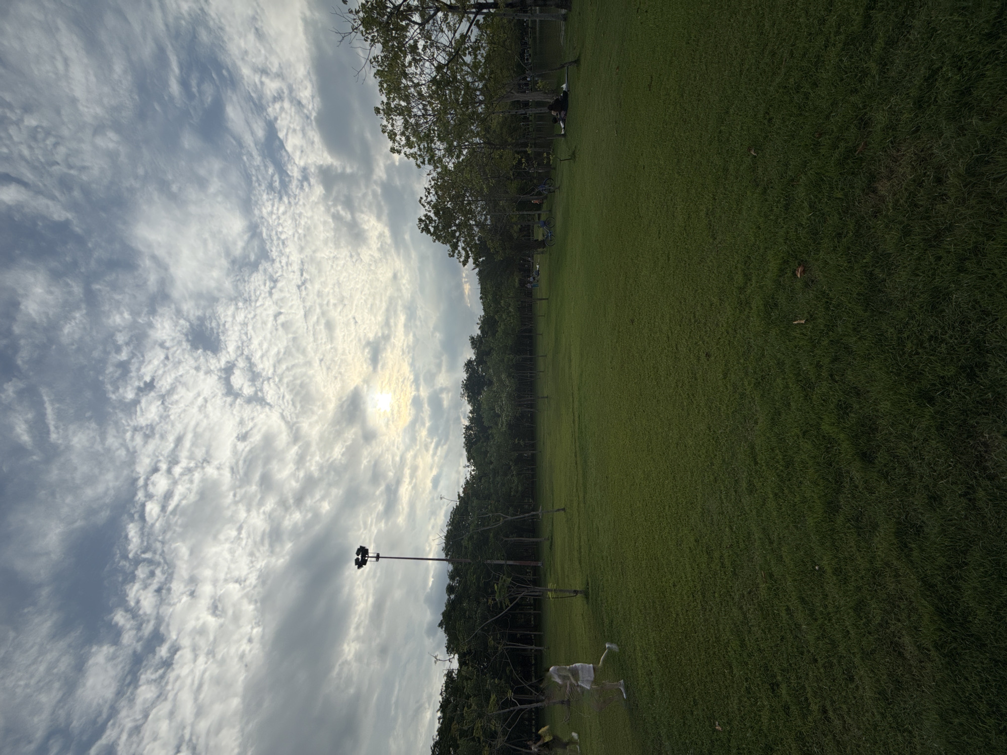 The image depicts a wide, grassy field in Lumphini Park, Bangkok, Thailand. The sky is overcast with dramatic clouds, and the sun is a bright disc partially obscured by the clouds, suggesting it is either sunrise or sunset. A tall lamppost stands prominently in the left half of the image, its lights pointing downwards. In the foreground, on the left, a woman in a white dress and sneakers is walking, her form slightly blurred, indicating motion. Further into the park, several trees form a dense line, and a body of water is visible in the distance on the right, with people sitting and relaxing by its edge. Two bicycles are parked on the grass in the middle ground. The overall atmosphere is serene, with a sense of people enjoying the open space.