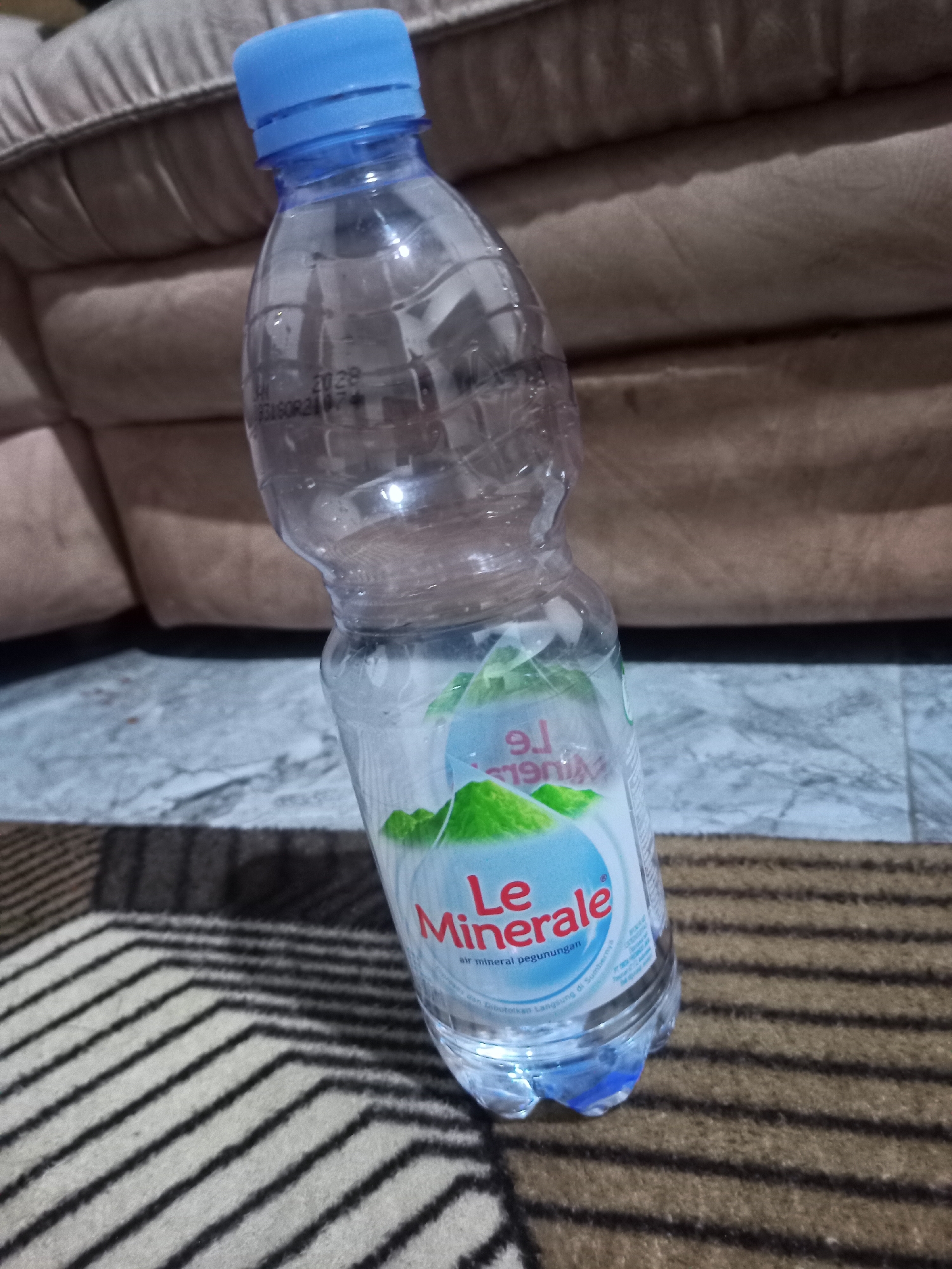 A partially filled bottle of Le Minerale bottled water stands upright on a striped carpet in what appears to be an indoor setting. The bottle has a blue cap and a clear plastic body. The label on the bottle is white with green mountain graphics and red text that reads "Le Minerale." Below that, in smaller text, it says "air mineral pegunungan" and "Dan Dibutkan Langsung di Sumbernya". The background consists of a beige sofa and a grey marbled floor. The lighting suggests it is daytime, likely indoors with artificial light. The image quality is somewhat blurry, particularly on the background elements. The presence of a carpet and sofa suggests a domestic or public indoor space. The specific location in the city of Karangtalun, Indonesia cannot be determined from the visual information provided in the image.