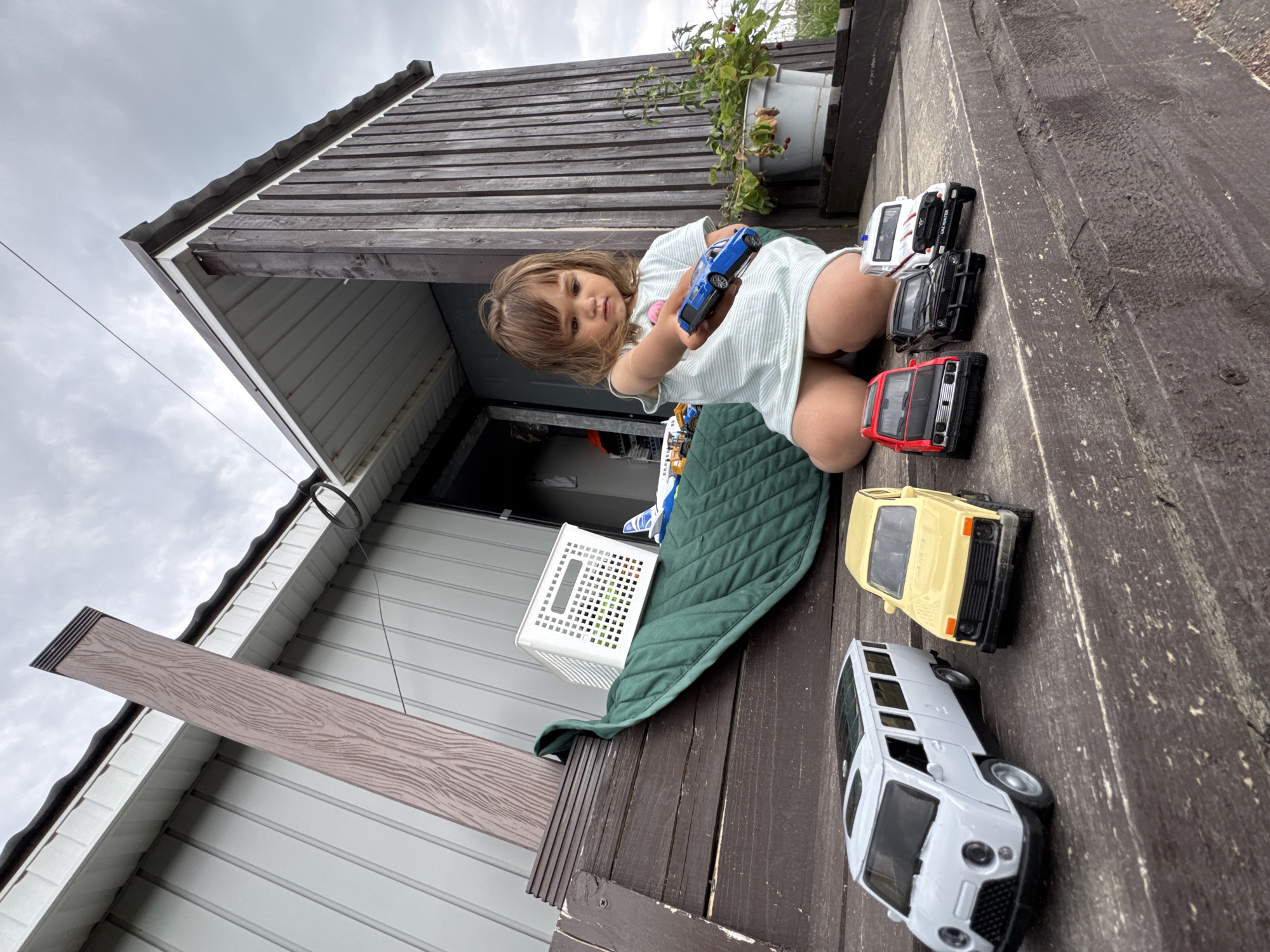 A young Caucasian female child, appearing to be around 2-3 years old, is kneeling on a wooden porch and playing with toy cars. She is wearing a light-colored, horizontally striped t-shirt. In her right hand, she holds a small blue toy car. In front of her on the porch are several other toy vehicles: a white van, a yellow SUV, a red hatchback, a black SUV, and a white vehicle with "UAZ HUNTER" written on it. The porch is made of dark wooden planks. Behind the child, a green quilted mat is spread out, with a white plastic basket placed on top of it. Further behind, a light-colored building with a corrugated metal roof is visible, along with a vertical wooden support structure that has a wood grain pattern. The sky is overcast with gray clouds. A potted plant with red berries is situated to the right of the child. The overall scene suggests an outdoor play area. The location is identified as Kempelevo, Russia.