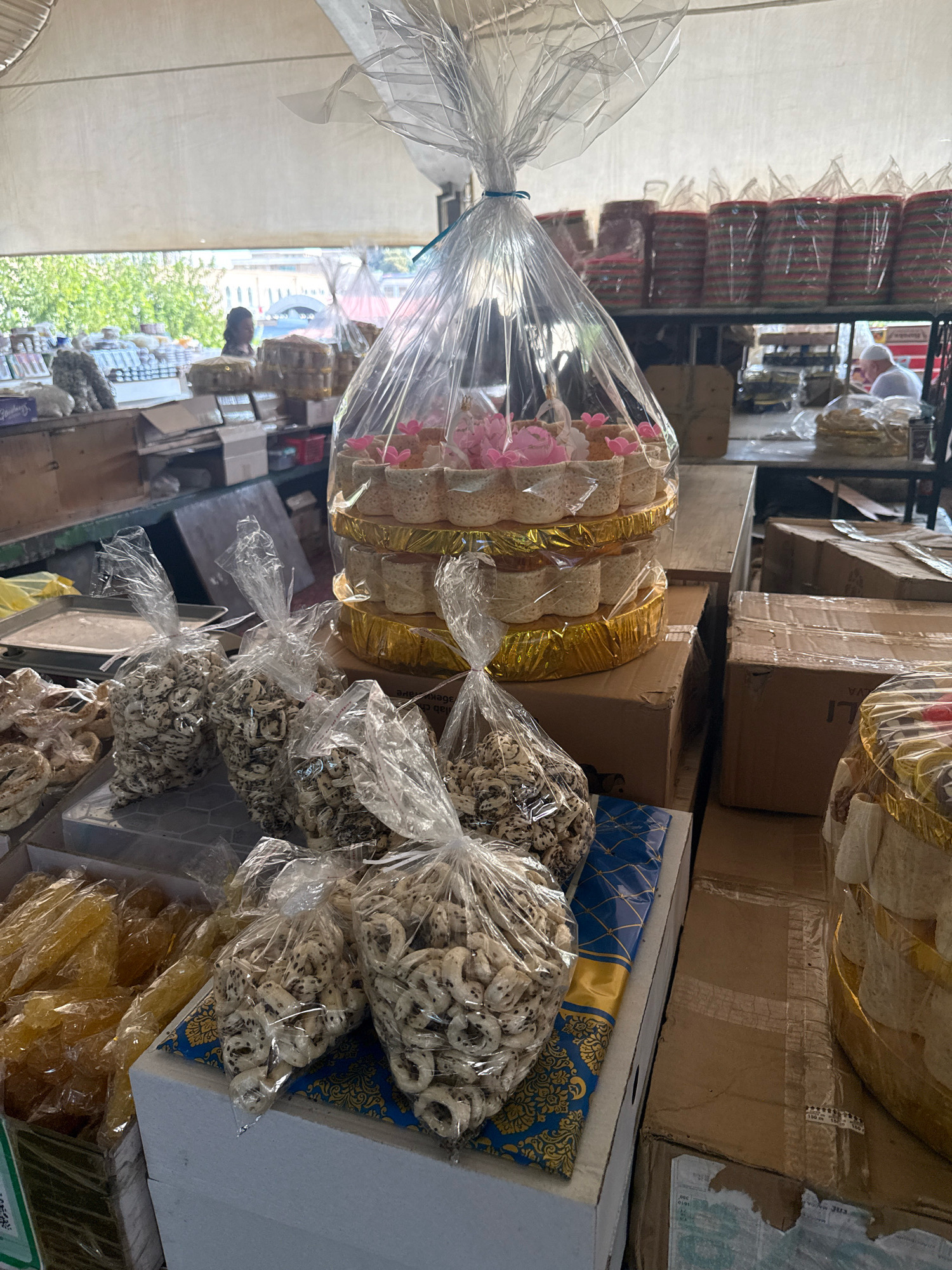 An indoor market scene shows a display of packaged goods. In the foreground, several plastic bags contain light-colored, ring-shaped snacks. Behind these, a multi-tiered cake or dessert display is wrapped in clear plastic and adorned with pink floral decorations. Further back, shelves are stacked with various boxed and packaged items. The environment appears to be a market stall with shelves and tables laden with goods. The lighting suggests it is daytime.