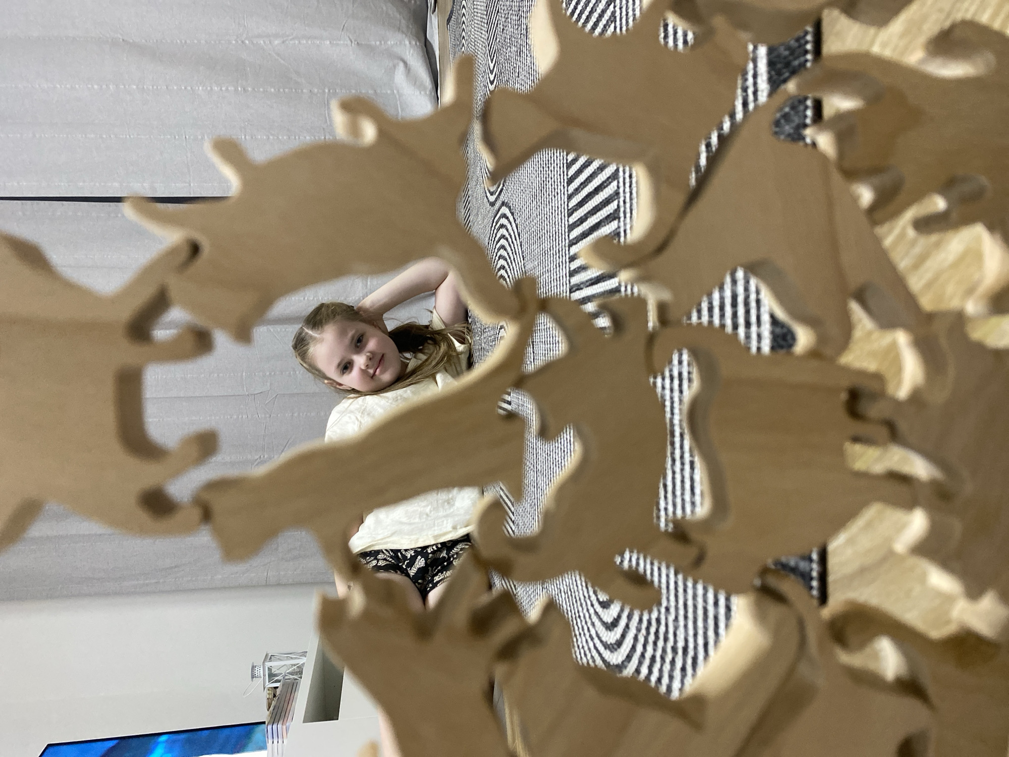 A young girl with blonde hair poses for a photo, viewed through a wooden animal-shaped toy. She is wearing a white blouse and floral shorts. The background includes a striped rug and a white wall. Part of a television set is visible in the lower left corner. The location and city of Izhevsk, Russia are not discernible from the image.