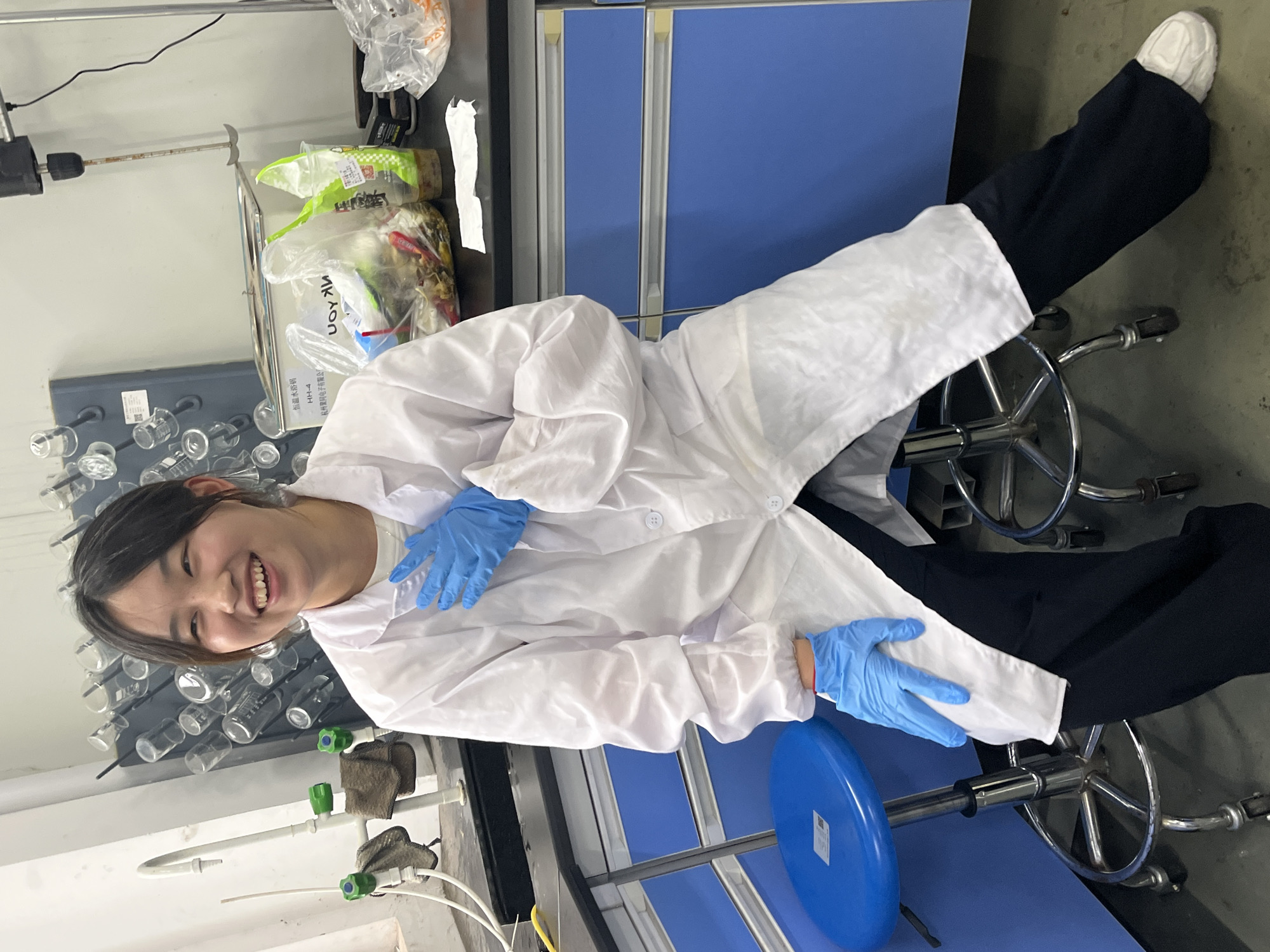 A smiling young woman wearing a white lab coat and blue gloves sits on a blue stool in a laboratory. She is indoors, possibly in a scientific setting. Behind her are lab equipment, including glassware and gas valves, indicating a chemistry or biology lab. In the background, there are blue cabinets. The location is Jiangjin, China.
