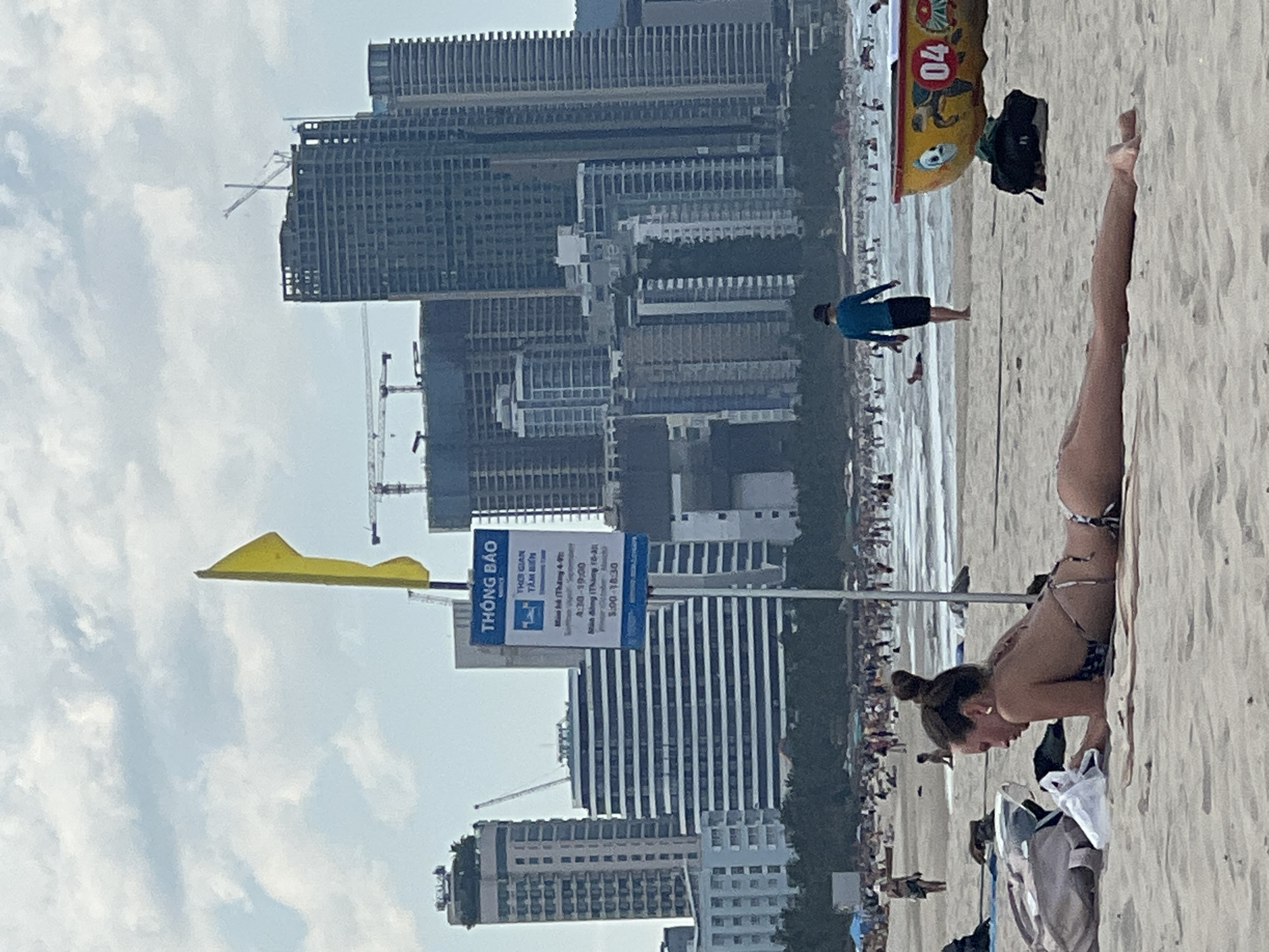 The image depicts a beach scene in Đà Nẵng, Vietnam, with a backdrop of modern high-rise buildings under construction. In the foreground, a young woman is lying on the sand, sunbathing. She is wearing a bikini and her hair is tied up in a bun. Beside her are beach accessories, including a bag. Further down the beach, other beachgoers are visible, some in the water and others walking along the shore. A yellow boat is partially visible on the right side of the frame. A blue sign with Vietnamese text and a yellow flag are also present. The sky is partly cloudy.
