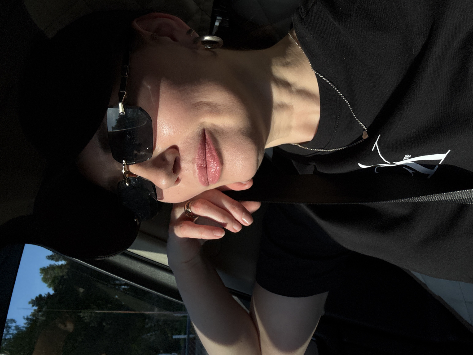 A person with fair skin is in a vehicle, wearing black sunglasses and a black t-shirt with a light-colored design on the chest, along with a thin silver necklace. The person is wearing a gold ring on one of their fingers. A black seatbelt is visible across their chest. The setting appears to be a car interior, with the window visible, showing trees and a blue sky. There is no information that can confirm that the image was taken in the city of Noviy, Russia.
