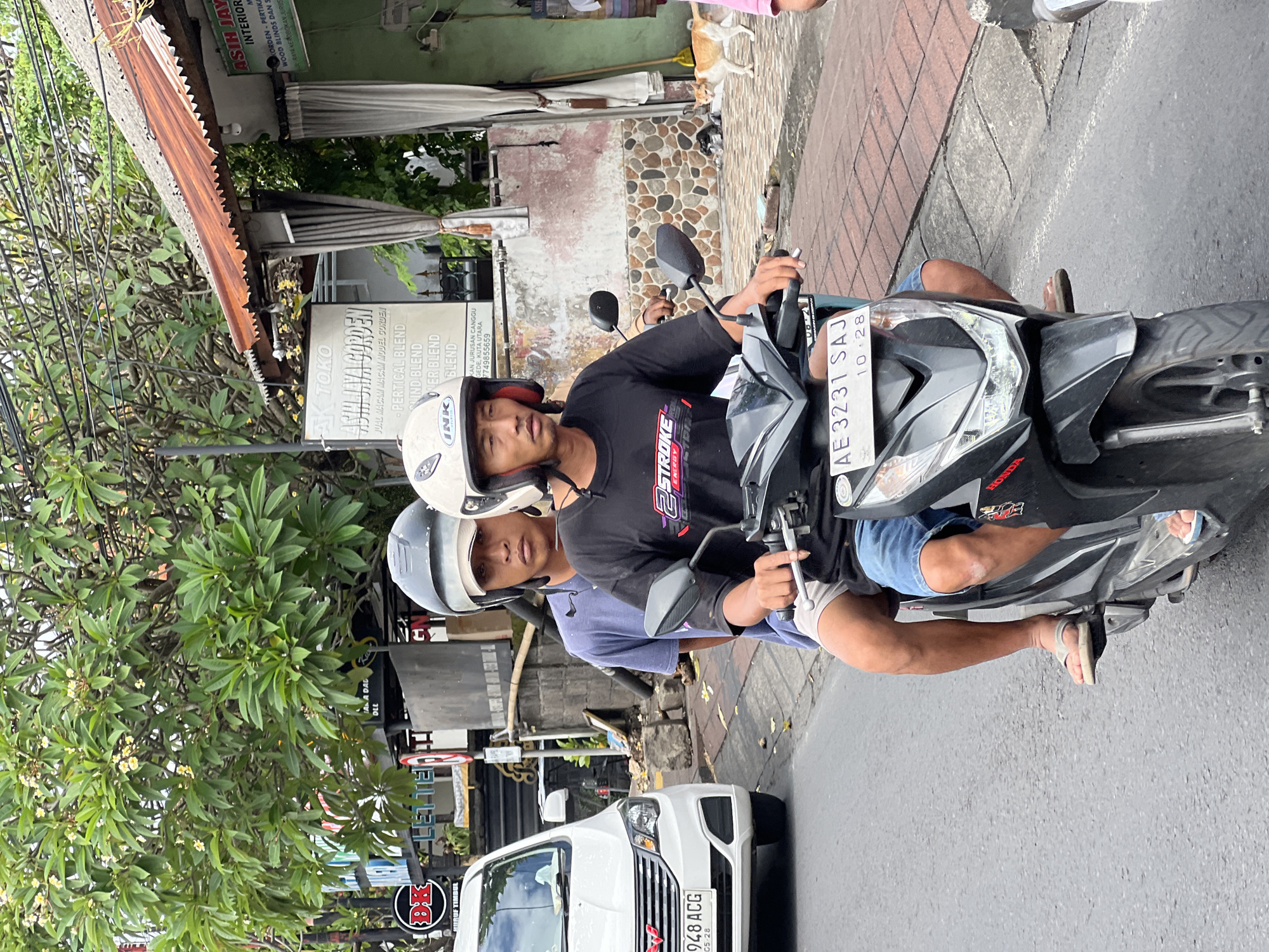 Two young men, wearing helmets, ride on a dark-colored scooter on a paved road in North Kuta, Indonesia. The man in front is driving, wearing a black t-shirt with red and white lettering, and blue shorts. The man behind him is wearing a blue t-shirt and white shorts. A white car with license plate "948 ACG" is visible to the left of the scooter, and a building with a corrugated metal roof is in the background. There are trees and signs on the side of the road. A light-colored cat is walking on the sidewalk in the background. The scene appears to be a typical street view in a Southeast Asian city.