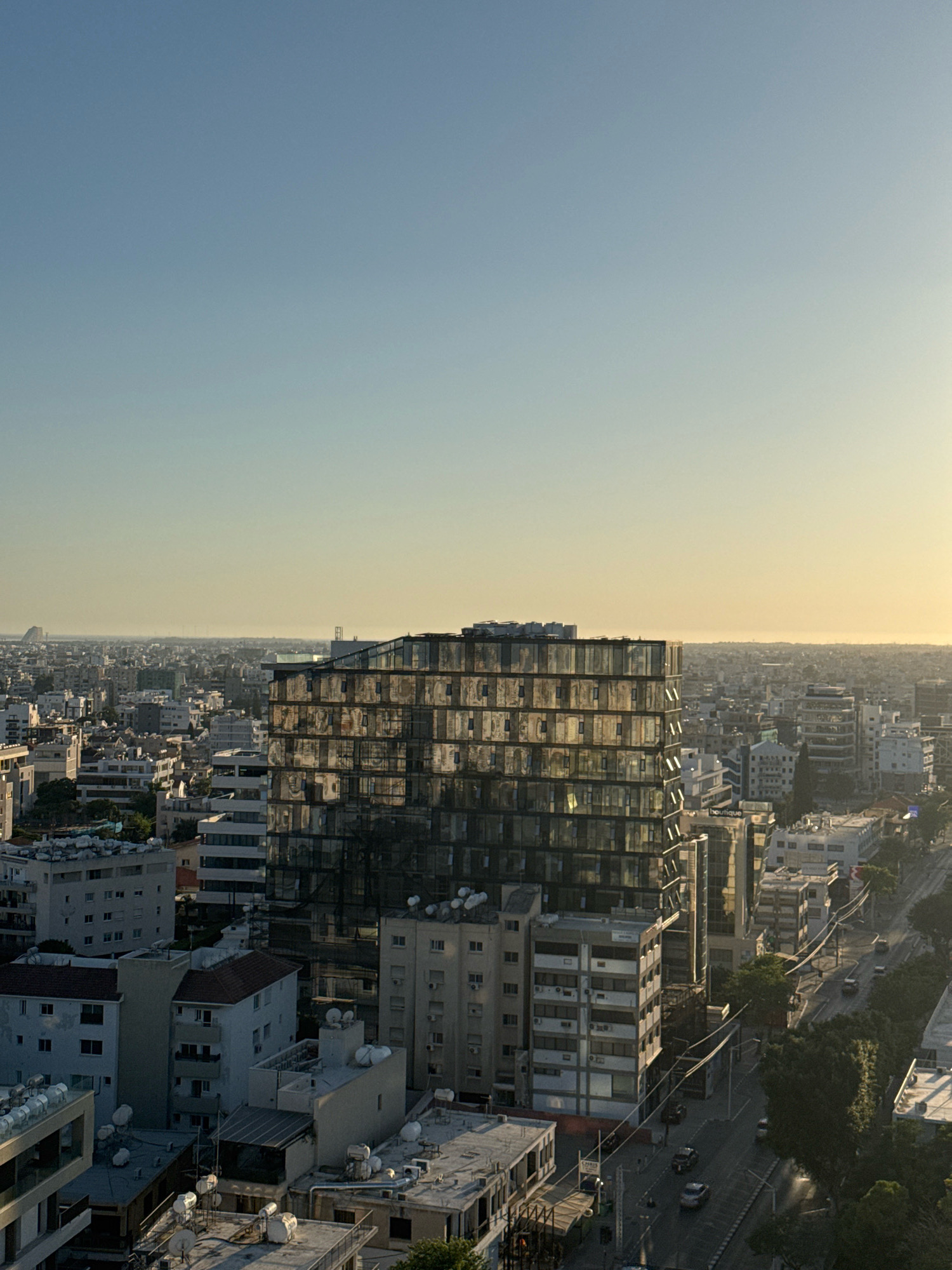 The image captures an aerial view of a cityscape, likely Limassol, Cyprus, bathed in the warm, golden light of either sunrise or sunset. The dominant feature in the foreground is a modern, multi-story building with a facade of reflective glass, creating a mosaic of mirrored windows. This building stands out against a backdrop of older, lower-rise structures, typical of urban development. Beyond the immediate buildings, the city stretches out towards a hazy horizon, where the sea or a similar body of water meets the sky. The atmosphere is serene, with the warm light casting long shadows and softening the edges of the urban landscape. The scene depicts a typical day in a Mediterranean city, with residential and commercial buildings coexisting. There are no discernible people in the image, and the focus is on the architectural and environmental elements of the city. The overall impression is one of urban density seen from a distance, with a sense of calm and natural light.