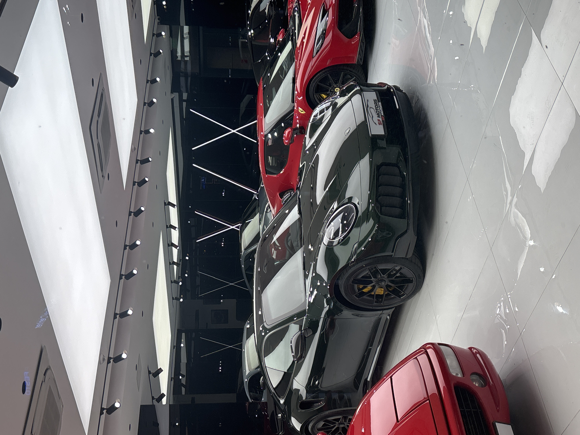 The image depicts a showroom in the city of Unknown, Unknown, displaying a collection of luxury cars. Prominently featured are a dark green Porsche, and multiple red cars of an indistinguishable brand. The cars are parked on a white tiled floor, which reflects light. The background includes a dark wall with illuminated linear designs and recessed lighting. The showroom has a modern architectural design.
