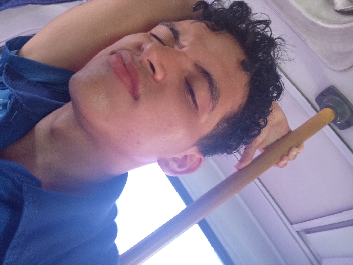 The image depicts a young adult male resting inside what appears to be a public transport vehicle, attributed to the city of La Plaza Paraguachí, Venezuela. The subject, a male with light skin, dark curly hair, and defined eyebrows, has his eyes closed. His head is tilted to the left, resting on his bent left arm, which is positioned behind his head. He wears a dark blue shirt with visible buttons. His right hand is holding a horizontal, yellowed or light brown grab bar, which is affixed to a light-colored overhead panel. A bright light source, consistent with a window, is visible in the lower background, creating a bright, somewhat overexposed area. The overall scene suggests the male is asleep or deeply relaxed while traveling.