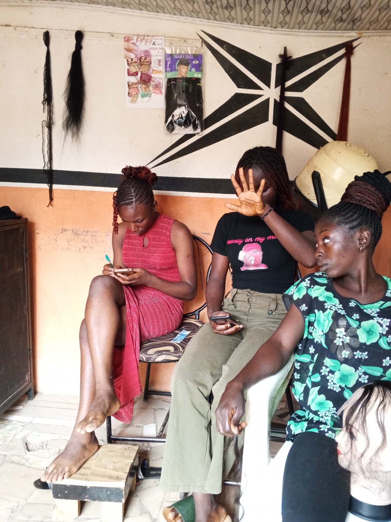 Three women are seated indoors in what appears to be a hair salon. The woman on the left wears a red dress and is looking at and writing on her phone. She has bare feet, with one resting on a small wooden footstool. The woman in the middle wears a black t-shirt with a pink graphic, and olive green pants. Her hand is raised, obscuring part of her face. The woman on the right wears a floral print blouse and black leggings. She is looking to the left and has a dark hairstyle and gold hoop earrings. A mannequin head with dark hair rests against her leg.
Various items are visible on the wall behind the women: two bundles of long black hair extensions, hair product advertisements, and a black and white geometric design painted on the wall. A hair dryer stands to the right. A dark cabinet is on the left. Based on the architectural features and style of the environment, the location is likely in Gwong, Nigeria.