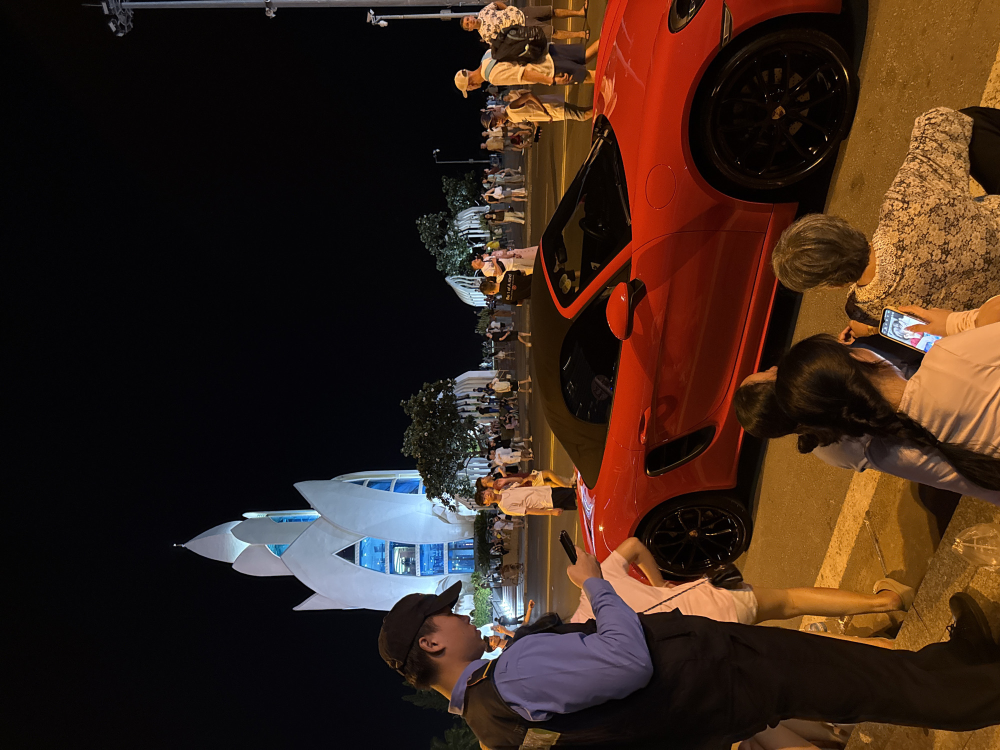 This is a night photograph taken in Nha Trang, Vietnam, near a distinctive white building with a lotus-shaped roof, likely a cultural landmark or religious site. In the foreground, a security guard in a dark vest over a blue shirt and a black baseball cap is looking at his phone. To his right, a vibrant red convertible sports car is parked. Several people are gathered around, with some seated on a curb in the foreground, including a woman with a patterned shirt looking at a phone and a young woman with braided hair. In the background, a crowd of people are walking and standing on a street and sidewalk, with trees and the illuminated building providing a backdrop. A tall light pole with multiple floodlights is visible on the right side of the image. The overall scene depicts a public gathering at night, possibly a tourist area or a cultural event, with the car and the unique architecture as prominent elements.