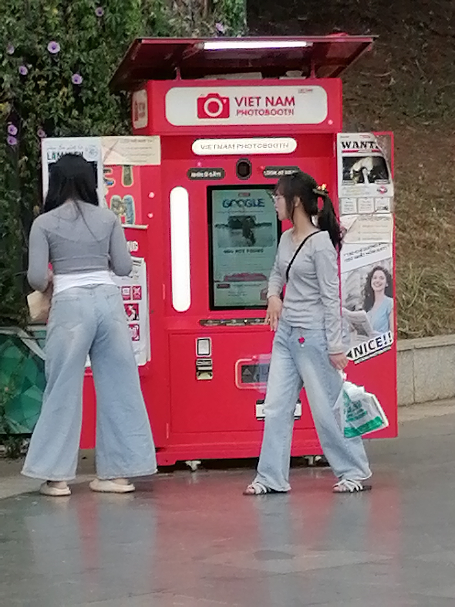 In Da Lat, Vietnam, a red photobooth machine is located outdoors. The machine features a large red frame with a glass display screen showing "GOOGLE." Above the screen, the words "VIET NAM PHOTOBOOTH" are displayed in white letters against a red background with a camera icon. To the right of the photobooth, a poster with the word "WANT" is attached. Two young women are standing in front of the photobooth. The woman on the left, viewed from the back, is wearing a grey cropped long-sleeved shirt and wide-leg light blue jeans. The woman on the right, viewed from the side, is wearing a similar grey long-sleeved shirt and wide-leg light blue jeans. She is holding a white plastic bag with green writing in her right hand and has a black strap across her chest. The ground in front of the photobooth appears to be wet, possibly from rain, and reflects the red color of the machine. Green foliage with purple flowers is visible behind the photobooth on the left.