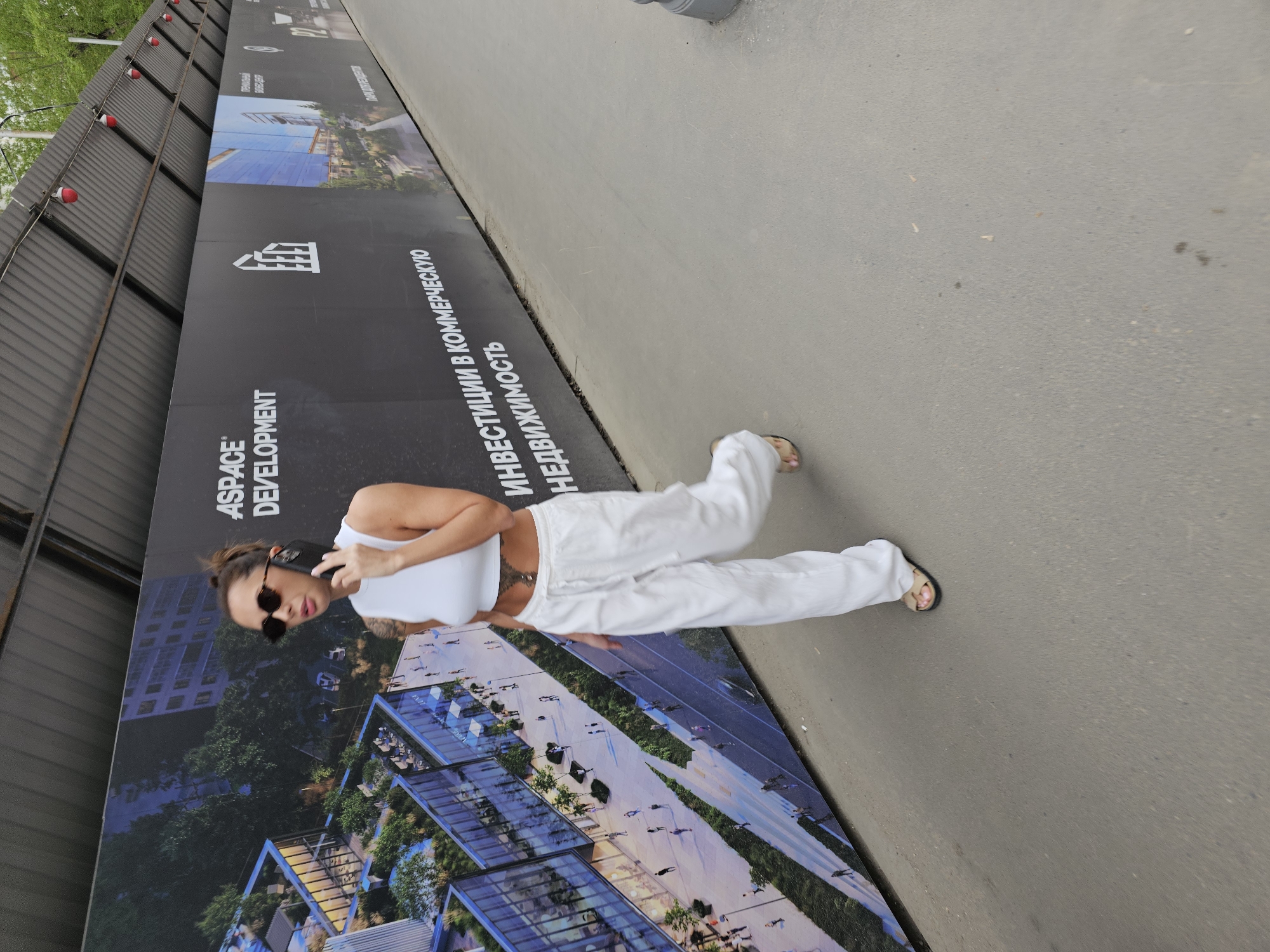 A woman with dark hair pulled up, wearing sunglasses, a white crop top, and white pants, walks along a sidewalk next to a construction barrier in Moscow, Russia. She is holding a cell phone to her left ear. The construction barrier is black and displays advertisements for ASPACE Development, with text in Russian that includes the phrase "инвестиции в коммерческую недвижимость" (investments in commercial real estate), along with images of architectural renderings. The sidewalk is made of gray concrete.