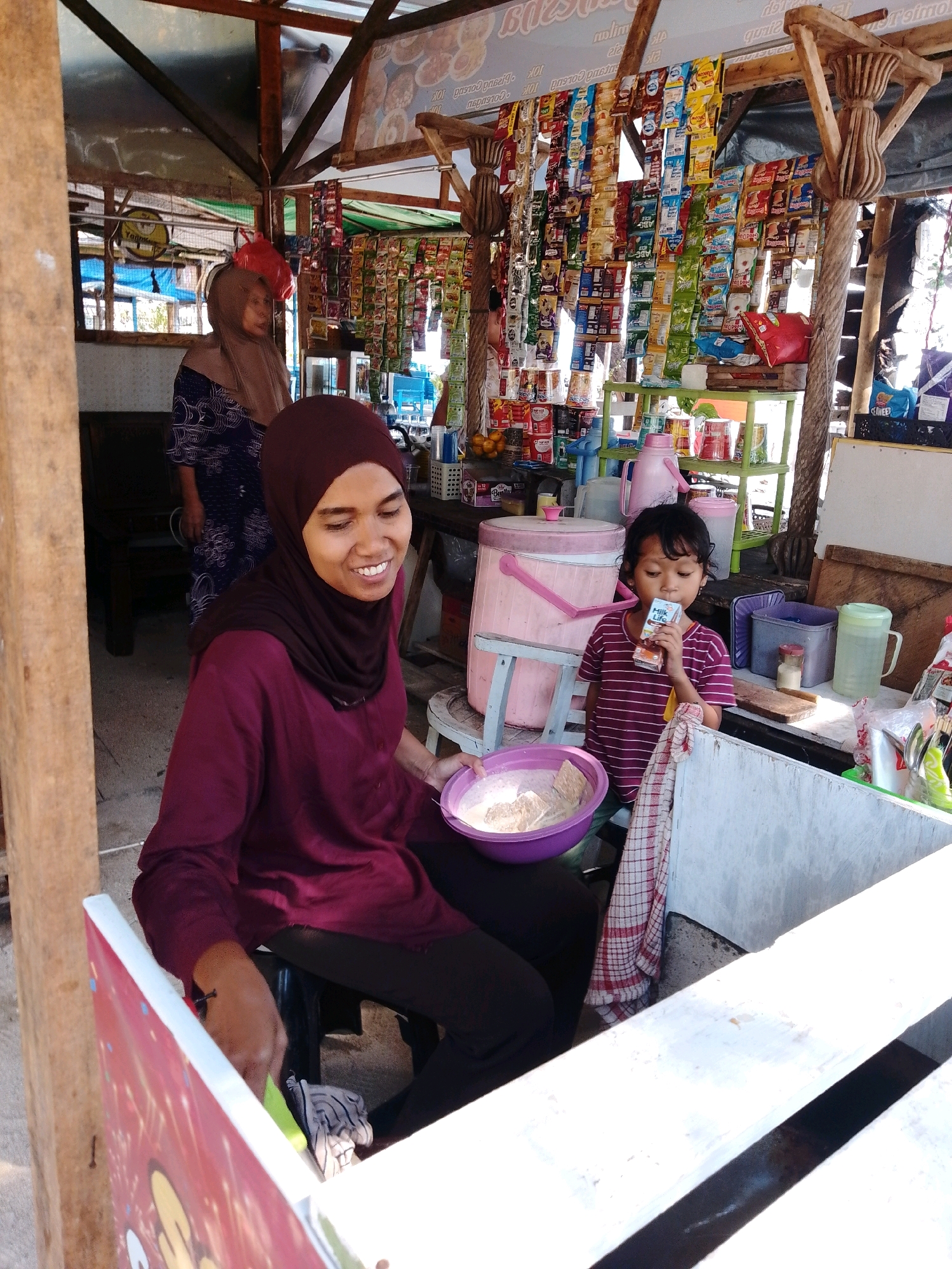 In Cikal, Indonesia, an outdoor market stall is depicted. A woman wearing a maroon headscarf and matching long-sleeved shirt is seated, holding a purple bowl containing a white substance, possibly food or ingredients. Her smile suggests she is engaged in a task or conversation. To her right, a young girl in a striped t-shirt is drinking from a small carton. Behind them, another woman in a headscarf and patterned dress stands near shelves stocked with various packaged goods, including snacks and beverages. The stall is constructed with wood and covered by a corrugated metal roof. Bags of merchandise hang from the ceiling. The overall scene captures a moment of daily commerce and interaction in a local Indonesian marketplace.