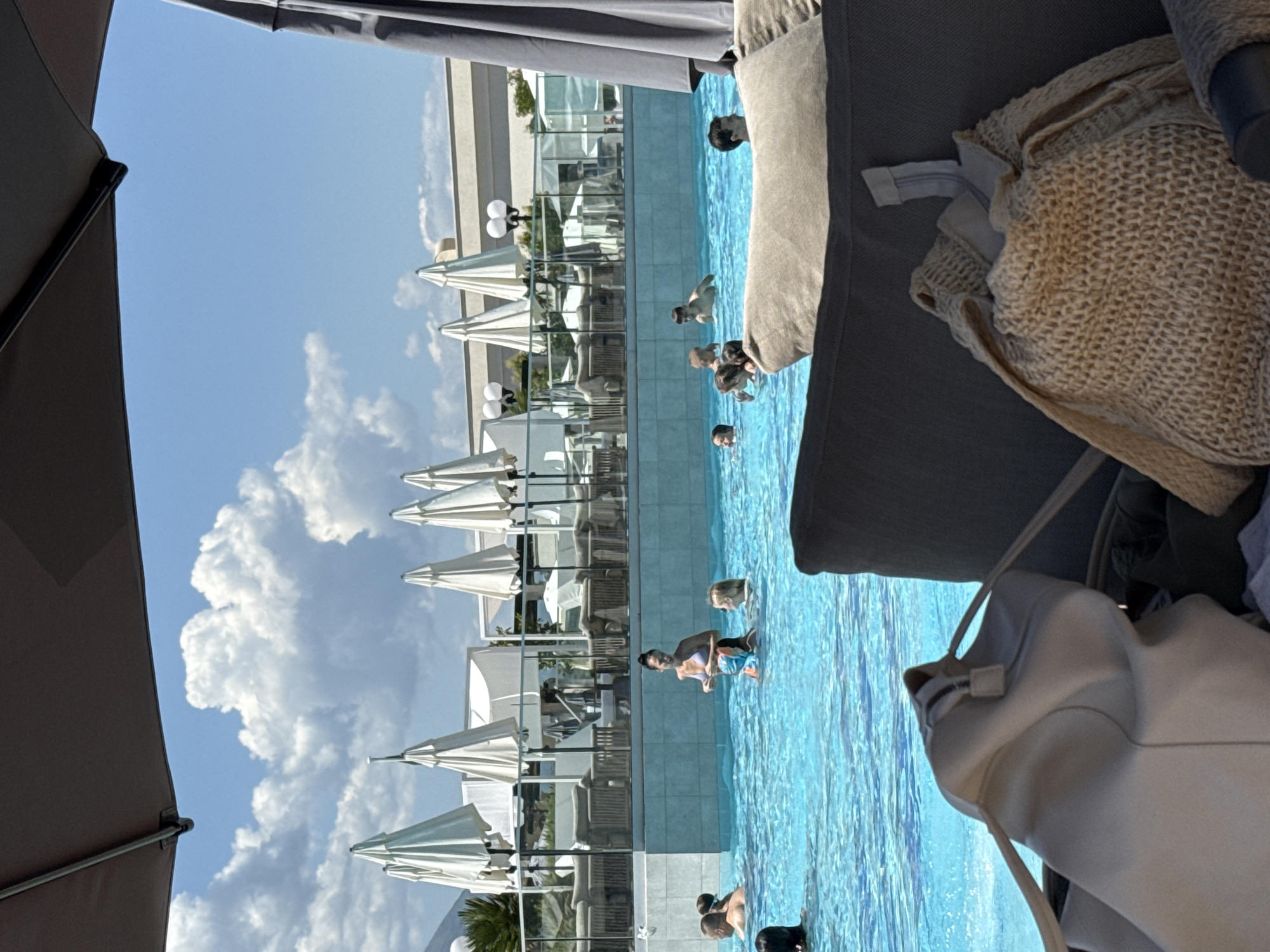 The image depicts an outdoor swimming pool area at a resort or hotel, location Unknown, Unknown, observed from a shaded seating position. A dark grey umbrella canopy is visible in the upper and right portions of the frame. In the immediate foreground, a dark grey cushioned lounge chair holds two bags: a light beige leather-like shoulder bag and a light brown woven net-style bag.
The central focus is a large, bright blue swimming pool containing several individuals. A woman wearing a light-colored bikini top stands in the pool, appearing to interact with a child or small person clad in colorful swim shorts. Other adults and children are partially submerged in the water. Behind the pool, a light-colored tiled wall supports a glass fence with dark railings, delineating a raised outdoor terrace.
On this terrace, multiple sets of dark grey cushioned outdoor sofas and tables are arranged under numerous large, white, conical outdoor umbrellas. Two street-style light fixtures with multiple white globes are visible on the terrace. In the background, a light-colored building with a flat roof is discernible, along with some green foliage. The sky is bright blue with several prominent white cumulus clouds. The scene suggests a sunny day.