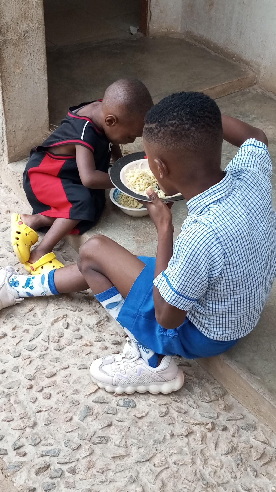 Two young African boys are sitting on a rough, stone surface in Sabon Tasha, Nigeria, sharing a meal of noodles. One boy is wearing a black and red tank top with yellow Crocs, and the other is in a blue plaid school uniform with white socks and sneakers. They are sharing noodles from a plate and a bowl. A concrete wall or pillar is visible in the background.