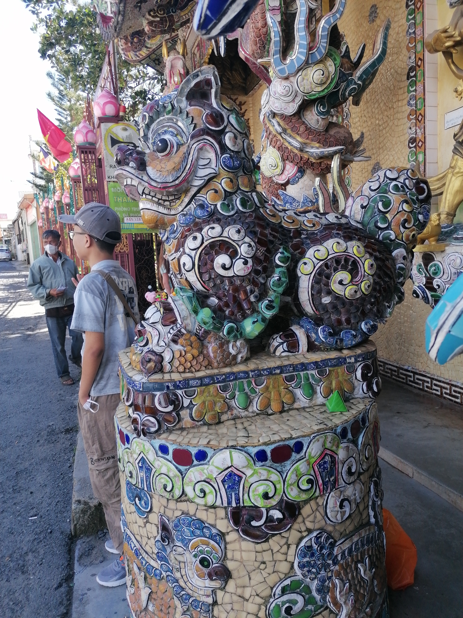 The image depicts an outdoor scene in Xuân Thành, Vietnam, likely a street or pathway leading to a temple or shrine, given the ornate mosaic decorations and religious imagery. In the foreground, a young person wearing a gray baseball cap, a light blue t-shirt, and khaki cargo pants stands facing right, holding a white mask in their hand. To the left of the young person, an older man wearing a light blue jacket, jeans, and a face mask is visible, holding a small object in his left hand. Dominating the center of the frame is a large, intricately decorated mosaic sculpture of a mythical creature, possibly a dragon or a lion. The sculpture is adorned with colorful ceramic tiles in shades of blue, green, red, white, and yellow, depicting swirling patterns and stylized animal features. The sculpture is mounted on a circular base, also covered in mosaics, featuring floral motifs and more creature imagery. Behind the sculpture, a building with a tiled facade and decorative elements is visible. In the background, a red flag and several smaller flags with Buddhist symbols are displayed. The ground is paved with asphalt. The overall impression is one of a vibrant cultural and religious site.