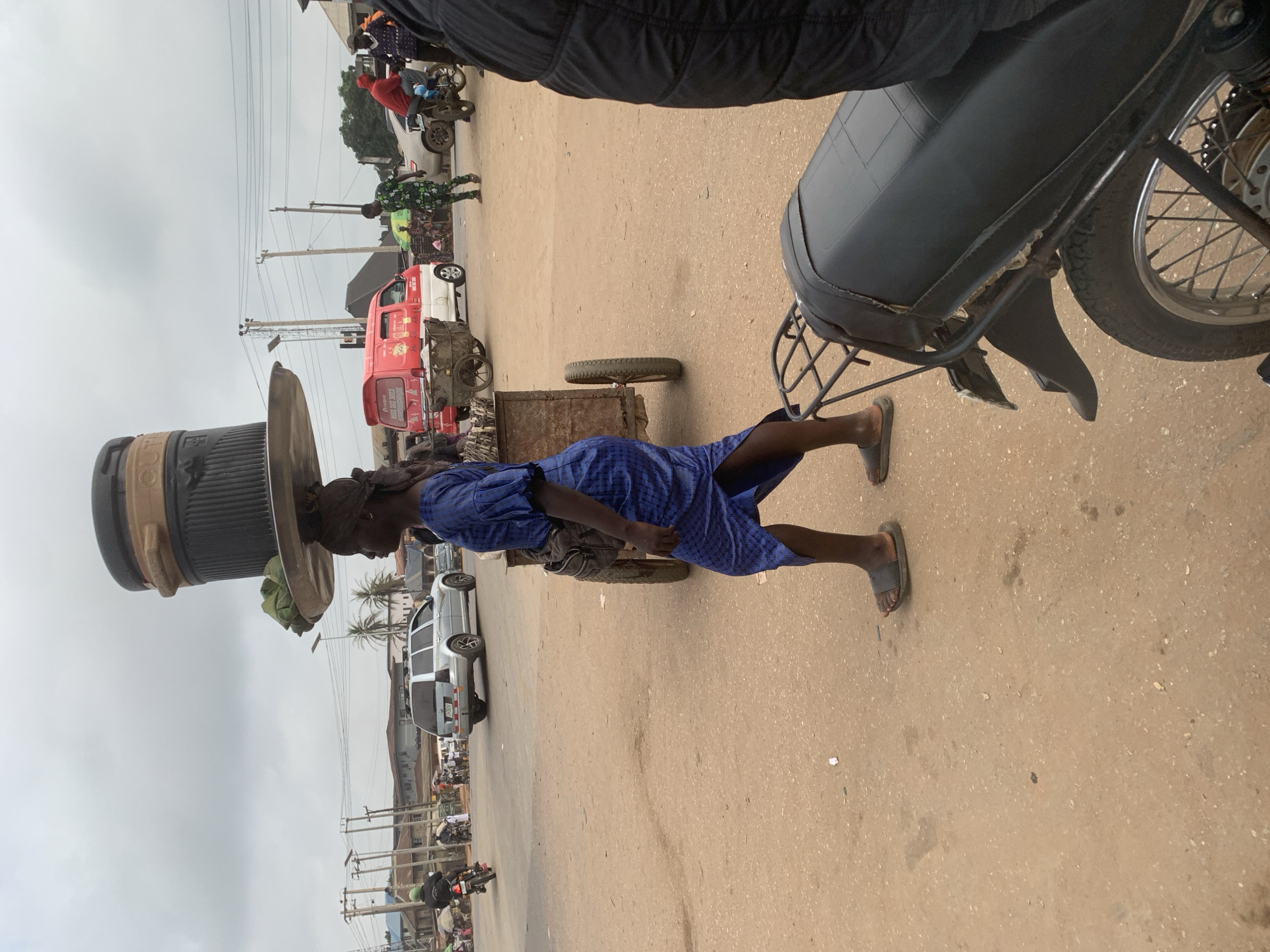 In Shagamu, Nigeria, a woman walks with a large container balanced on her head, supported by a metal tray. She wears a blue dress and sandals. To her right, a cart with two wheels is partially visible. In the background, a silver SUV is parked, and other vehicles, including a red and white van and motorcycles, are present. The scene appears to be a street or marketplace with overhead power lines and buildings lining the road, under a cloudy sky.