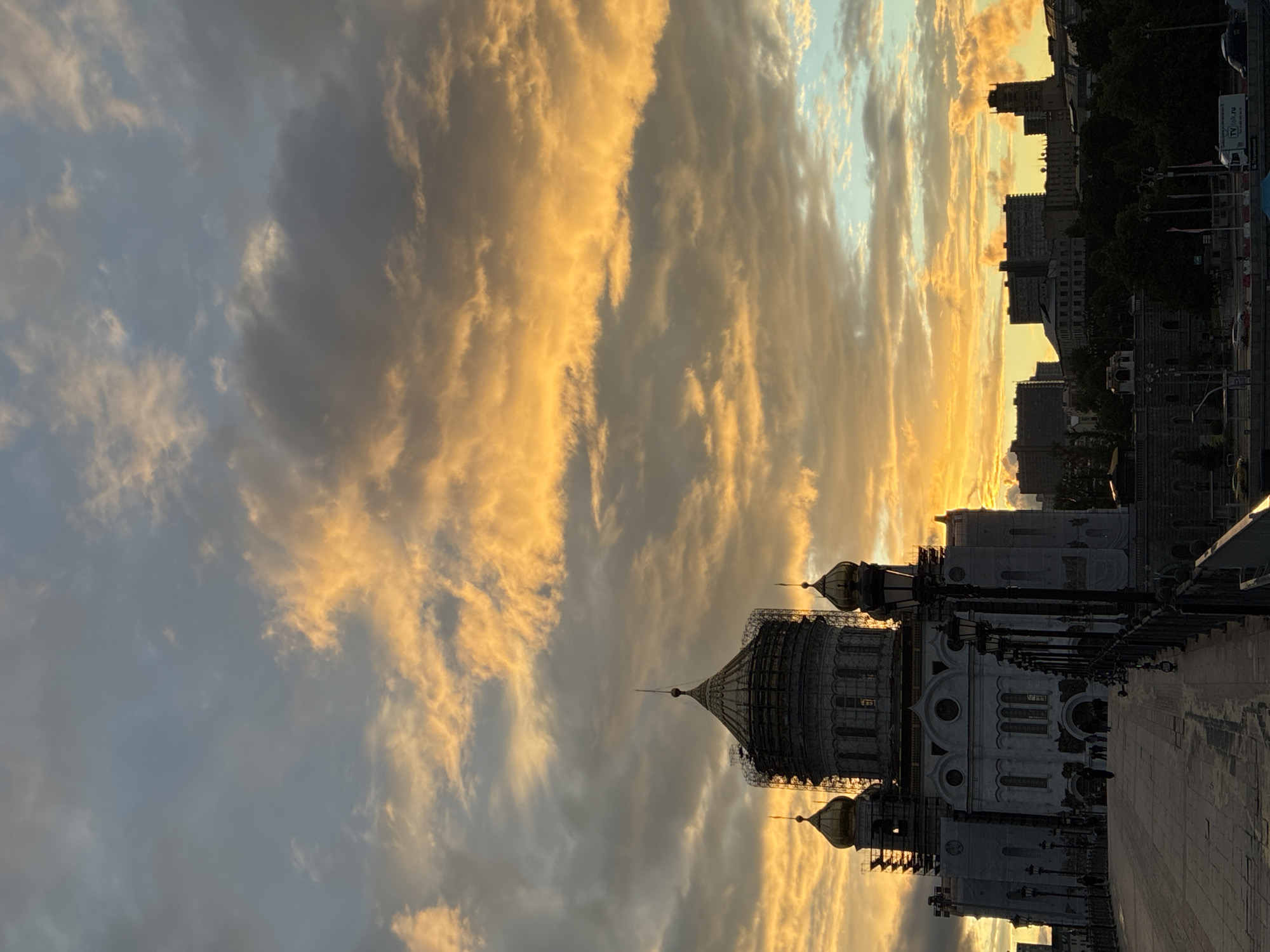 The image depicts the Cathedral of Christ the Saviour in Moscow, Russia, during a sunset. The sky is filled with dramatic, colorful clouds illuminated by the setting sun. Scaffolding is visible on parts of the cathedral, suggesting ongoing maintenance or restoration. Buildings and silhouettes of trees are visible in the background. A paved walkway is present in the foreground.