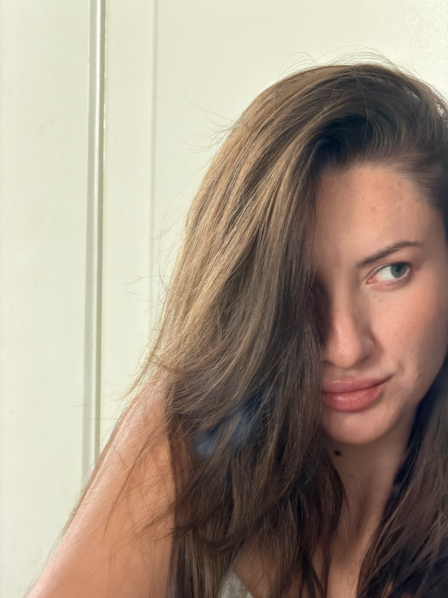 The image shows a close-up of a woman's face, with her hair obscuring part of her left cheek and eye. She has long, brown hair, and her right eye is visible, looking to the right. Her lips are slightly parted and appear to have a glossy finish. The background is a plain, light-colored wall with vertical lines, suggestive of a door or partition. The lighting is soft, creating a gentle illumination on her skin. The overall impression is of a casual, possibly indoor, setting. There is no indication of the location being Sharm El Sheikh, Egypt.