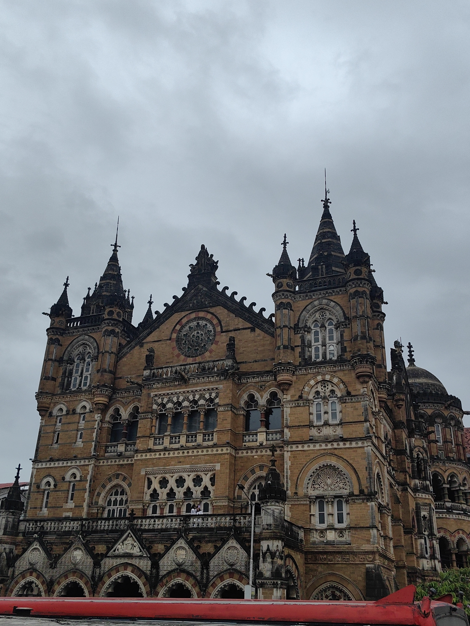 The image displays the Chhatrapati Shivaji Maharaj Terminus (CSMT), formerly Victoria Terminus, in Mumbai, India. This UNESCO World Heritage Site is a historic railway station renowned for its Victorian Gothic Revival architecture. The structure is made of sandstone and features ornate carvings, turrets, domes, and a large clock tower. The sky above is overcast and grey. A red-colored object, possibly the roof of a vehicle or train, is visible at the bottom of the frame, partially obscuring the base of the building. A few individuals can be seen standing on a balcony on the building's facade.