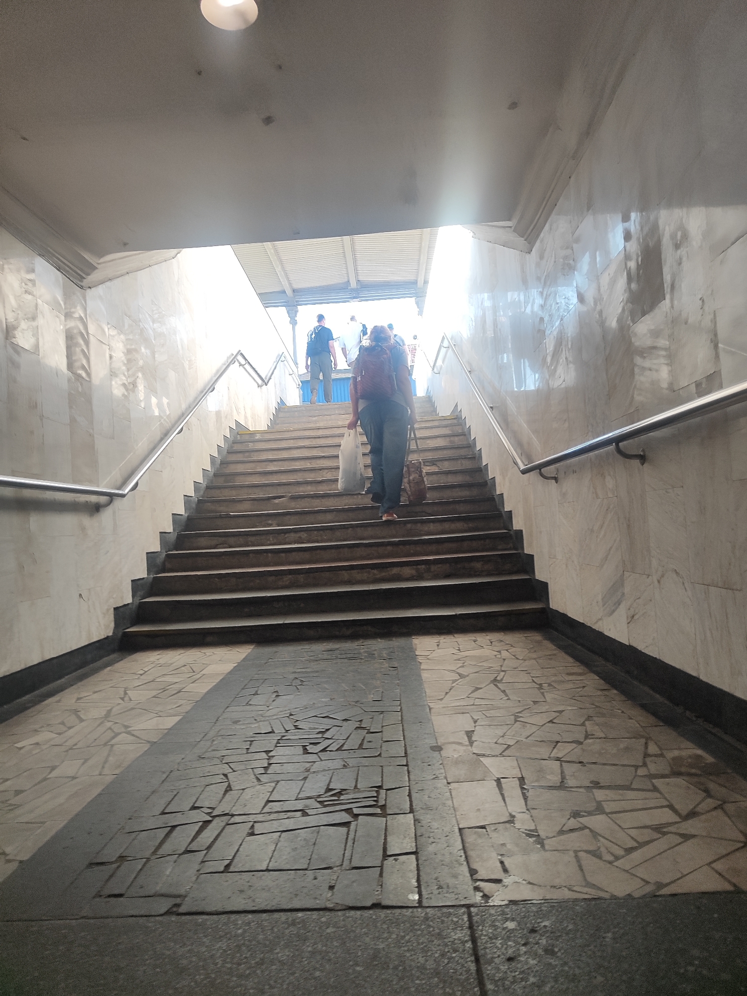 The image shows a wide, concrete staircase leading upwards in what appears to be a public underpass or transportation station. The walls on either side of the staircase are covered in light-colored marble tiles, which reflect the bright light coming from above. A metal handrail runs along the right side of the stairs.
Several people are ascending the stairs. In the foreground, on the right side, a person with a dark backpack and wearing dark pants is carrying a white plastic bag. Further up the stairs, two individuals are visible, one with a dark backpack and the other in a white shirt. The floor at the base of the stairs is paved with irregular, dark grey and light grey tiles, with a distinct shadow line dividing them. The ceiling above the stairs is a smooth, light-colored surface with a circular light fixture. The overall lighting suggests it is daytime, with a strong source of natural light at the top of the staircase.