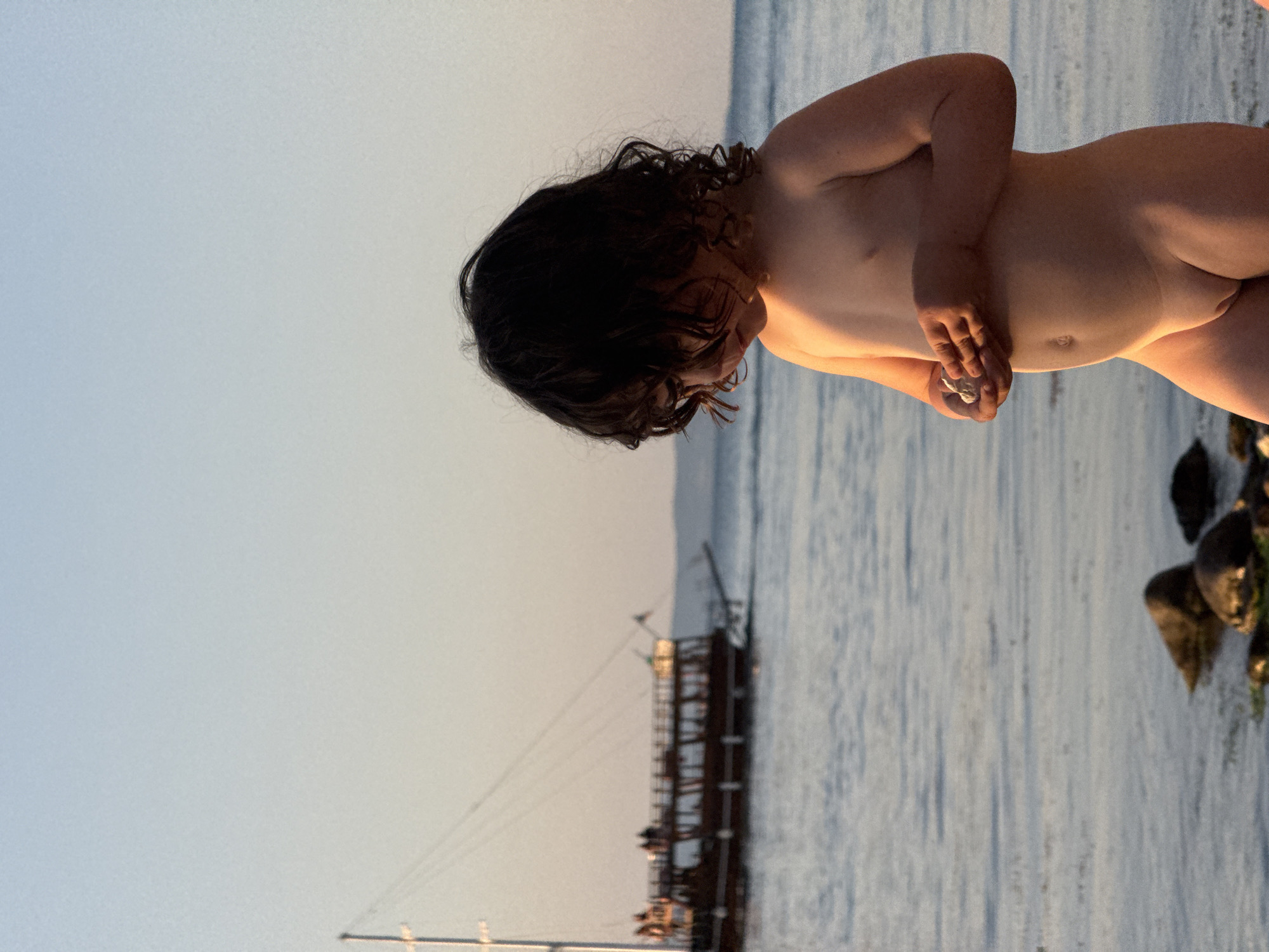 This is a photograph of a young child, likely a toddler, standing on the shore of a body of water. The child has dark, curly hair and is facing downwards, appearing to be looking at something in their hands. Their arms are crossed, and they are unclothed. The water in the background is calm and reflects the light, suggesting it's either sunrise or sunset. The sky is a pale blue, indicating clear weather. In the distance, a traditional wooden sailing ship with multiple decks and masts is visible, moored in the water. The ship's design and the overall serene atmosphere suggest a coastal location. The photograph's orientation is landscape, and the lighting suggests it was taken during the golden hour. The scene evokes a sense of peaceful contemplation.