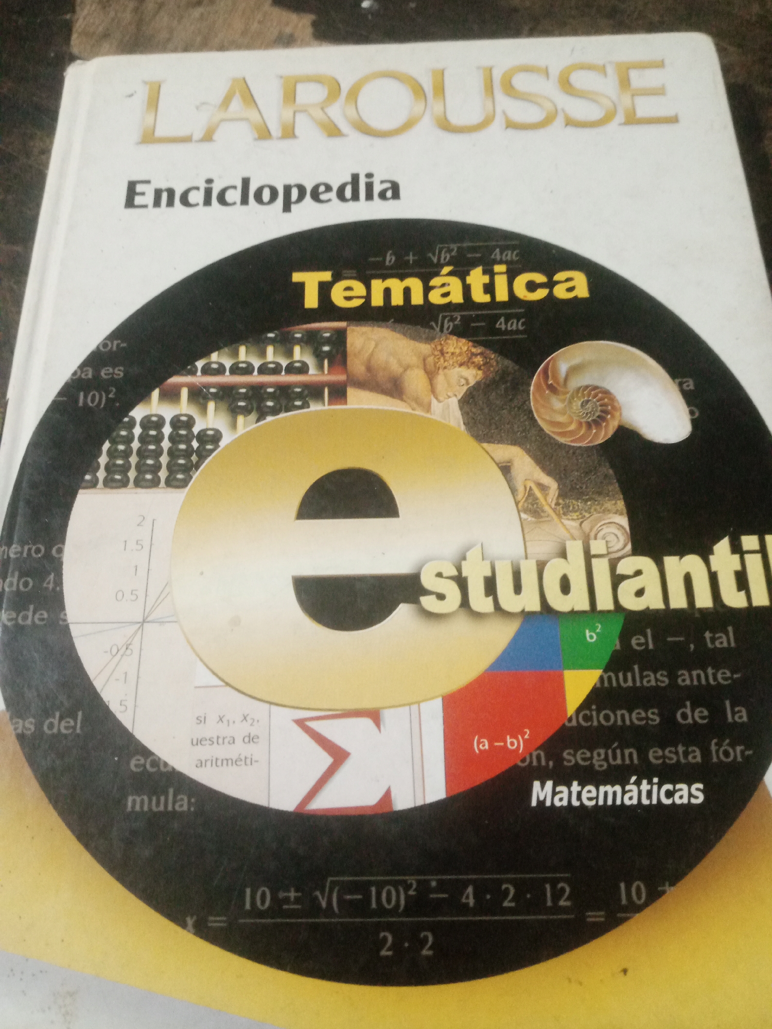 The image is a close-up, high-angle shot of the cover of a book titled "LAROUSSE Enciclopedia Temática". The book cover is mostly white with gold lettering for the title. A large, circular graphic is superimposed on the lower half of the cover. This graphic features a large, stylized "e" in gold, a nautilus shell, an abacus, and an image of a classical figure. Text on the graphic includes mathematical formulas and the words "estudiantil" and "Matemáticas". The lighting is somewhat uneven, with a strong glare on the upper portion of the book. The book is lying on a textured surface, possibly a table or wooden furniture. No people are visible in the image. There is no information within the image to suggest the location is Santa Ana, Venezuela.