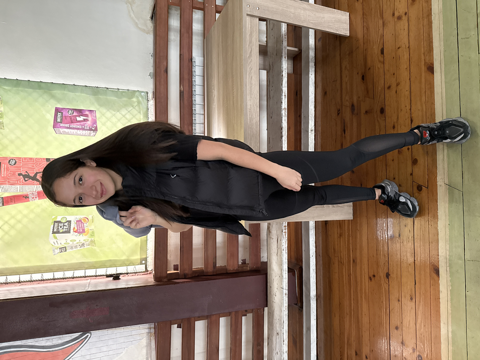 An individual, identified as a young female, is positioned centrally, standing in an indoor facility in Sofia, Bulgaria. She is facing slightly right of the camera, with a mild smile. She has long, dark brown hair. She wears a black puffer vest over a black short-sleeved top, black leggings with sheer panels on the lower legs, and black and grey athletic shoes with red accents. A light grey garment is draped over her left shoulder, held by her right hand.
The background consists of a glossy, amber-toned wooden floor in the foreground, transitioning to a lighter green-painted section and then exposed wooden planks. Behind the subject, the lower wall features multiple rows of dark brown wooden slats, likely tiered seating or benches. Above these slats, a white-painted wall section exhibits localized water damage and staining, particularly in the upper right. On this wall, two advertisements on a light green background are visible: one for "INSTICK ICE TEA" (The Launch Extra with 20mg Caffeine, Orange & Passion Fruit) and another for "INSTICK ENERGY DRINK" (with a lightning bolt graphic). A prominent, dark reddish-brown vertical structural beam is present on the left side, partially obscuring a red and white graphic. A light-toned wooden table with angular legs is positioned to the subject's right. A white radiator is visible behind the wooden slats in the background.
