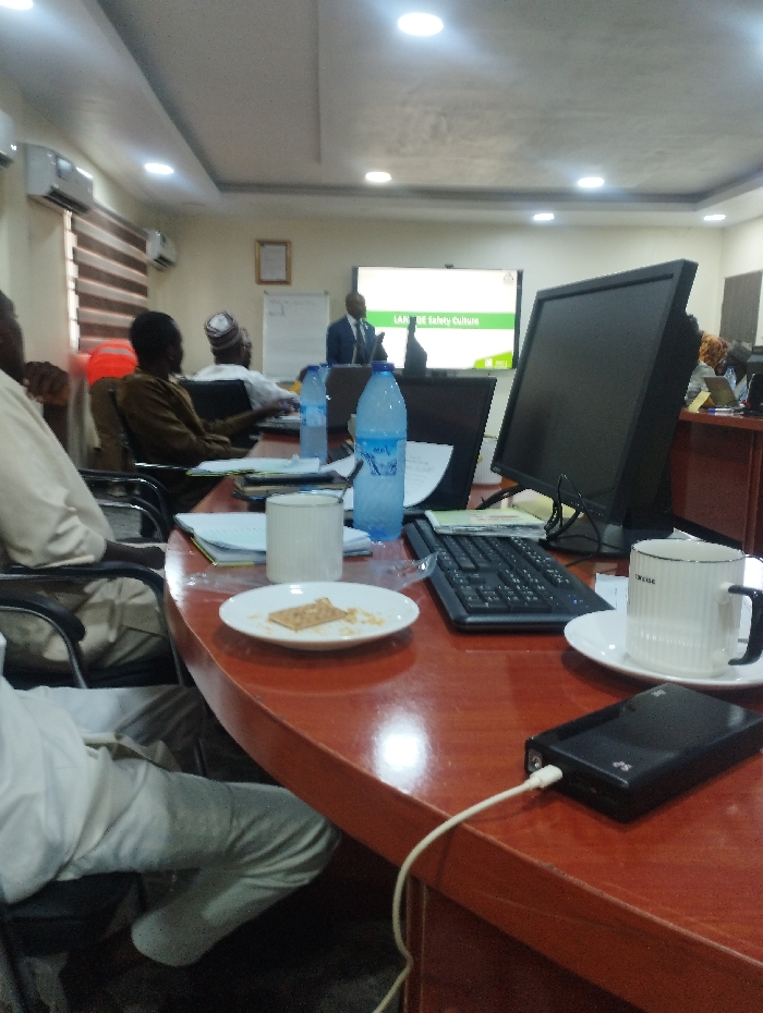 The image shows a conference room in Ashaka, Nigeria (10.929, 11.476). There are four men in the room, three of whom are sitting at a long wooden table. The man at the far end of the table is wearing a white shirt and white pants, while the man next to him is wearing a brown shirt. A third man is wearing a white shirt and a white cap. The fourth man is standing at a whiteboard behind the table, giving a presentation. The whiteboard reads "L.A.R.E Safety Culture". There are a few objects on the table in front of the man in the white shirt, including two clear water bottles, two coffee mugs, a plate with a piece of bread, a keyboard, a black laptop, a small black rectangular object, a white cable, and some papers.