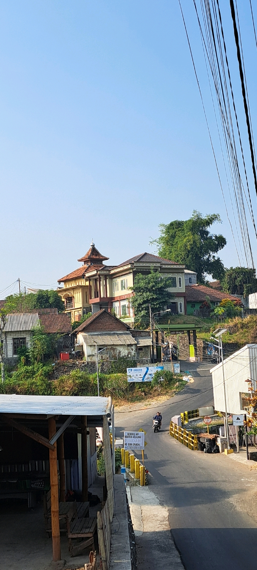 The image shows a street scene in Gambiran, Indonesia. In the foreground, there is a wooden structure with a corrugated metal roof. To the right, a paved road curves away, with a person on a motorcycle traveling on it. The road is lined with yellow barriers and a sign that reads "STOP". Further down the road, there is a white building with a dark-colored roof and a sign with "MOHAMMADTHROPI" written on it. In the background, several houses are visible on a hillside, with lush green vegetation surrounding them. The sky is clear and blue.