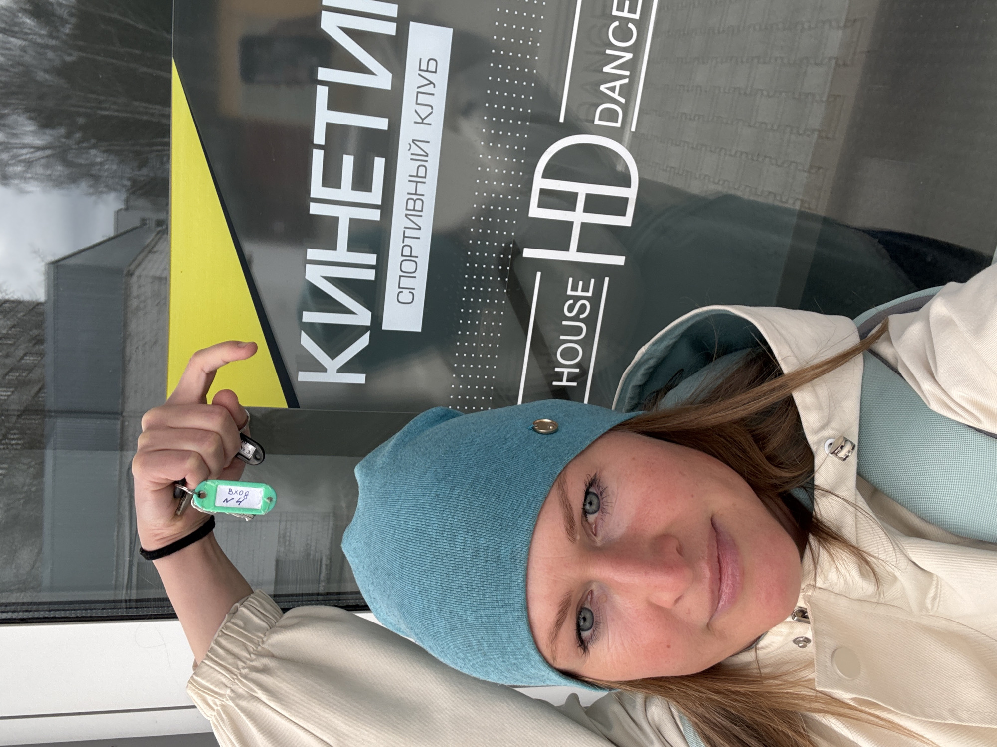 A woman is standing in front of the “KINETIKA SPORTIVNYI KLUB” in Chaikovsky, Russia, holding up a set of keys in her left hand. The keys have a green key tag labeled “Bxog N4”. She is wearing a light blue beanie and a beige jacket. The sign behind her advertises the KINETIKA SPORTIVNYI KLUB, a sports club, and HD House Dance. The sign is black and gray with a strip of yellow on the left edge. The background shows a building and trees in the distance. The woman appears to be taking a selfie.