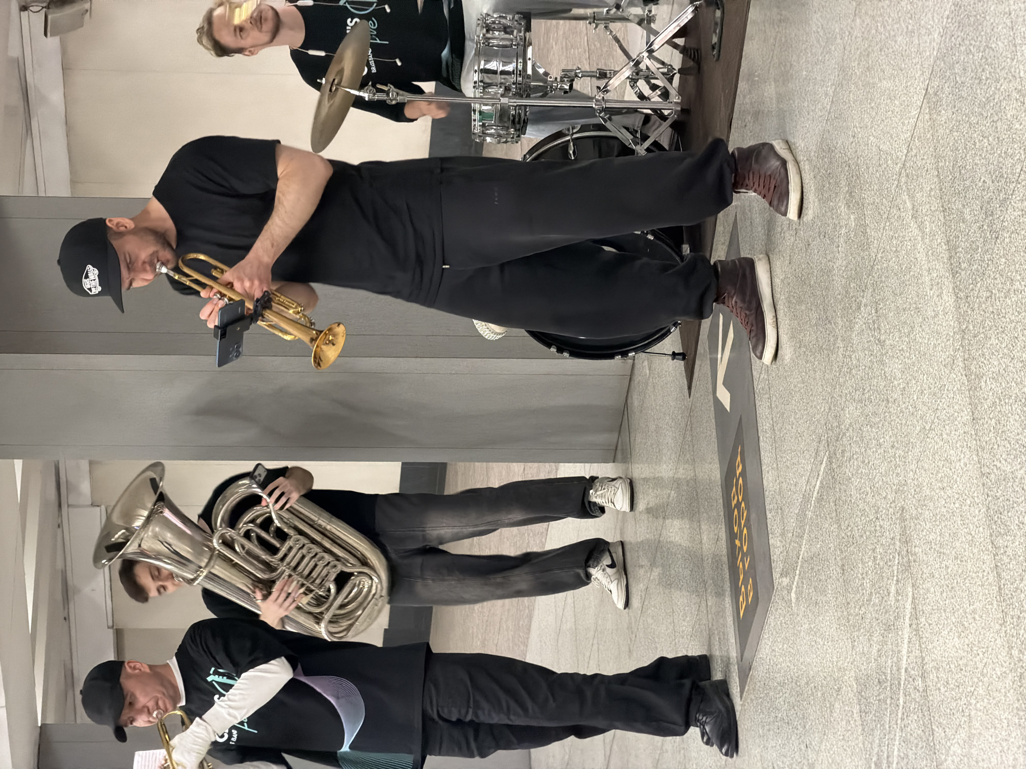 The image depicts a brass band performing in what appears to be a Moscow Metro station. Four men are visible, three of whom are actively playing instruments. The man on the far left is playing a trumpet, wearing a black t-shirt, black pants, and a black baseball cap. The second man from the left is holding a large tuba and wearing a black t-shirt and dark pants. The central figure is playing a trumpet, mounted on a stand, while wearing a black t-shirt, black sweatpants, and a black baseball cap. To the right, a fourth man is seated behind a drum kit, partially visible, wearing a black t-shirt with some white lettering. The environment features typical metro station flooring with visible tile patterns and a grey wall in the background. A sign on the floor in the foreground reads "ВЫХОД В ГОРОД" (Exit to the city) in yellow lettering.