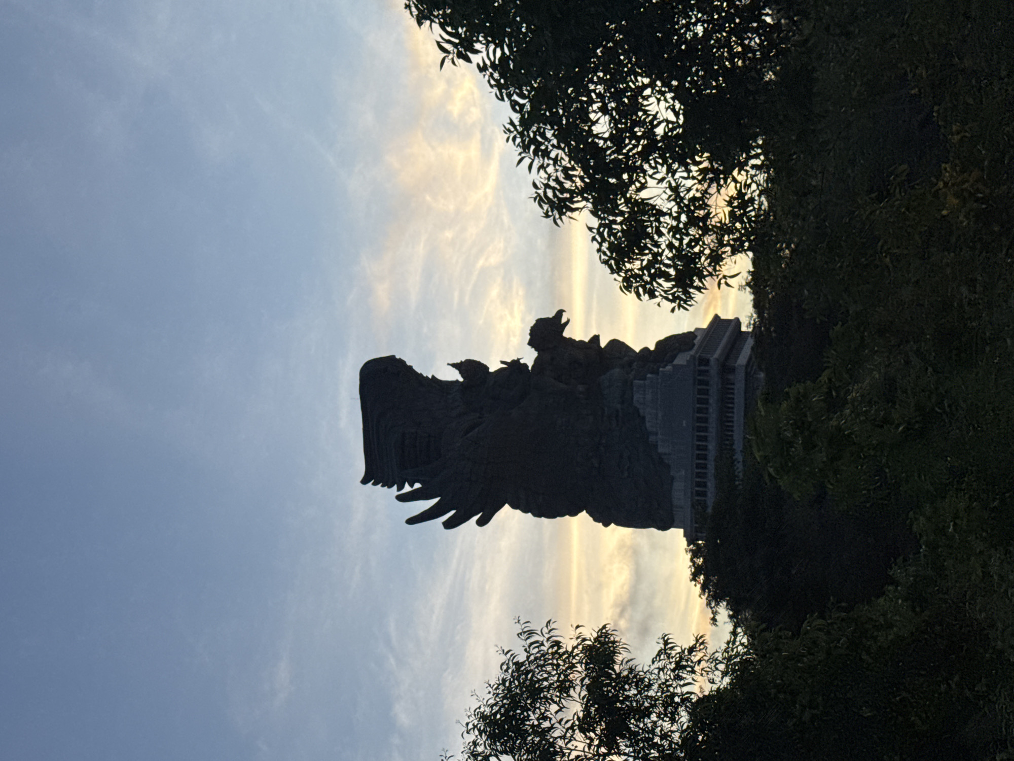 The image, oriented vertically, captures the majestic silhouette of a colossal statue against a dramatic twilight sky, framed by dark foliage. The central subject is a towering monument, likely the Garuda Wisnu Kencana statue, depicting a large, mythical bird-like creature – possibly Garuda – with a smaller, distinct figure, presumably the deity Wisnu, seated on its back. The statue is rendered in complete silhouette, showing its grand scale and intricate outline, including the prominent wings and detailed architectural base, which appears to have multiple tiers.
The sky occupies a significant portion of the background, displaying a gradient of soft blues at the top transitioning into warm golden and orange hues closer to the horizon, indicative of either sunrise or sunset. Wispy, scattered clouds are visible, catching the fading or rising light.
Dark, leafy trees and dense foliage frame the top and bottom right of the scene, also in deep silhouette, further emphasizing the vibrant sky and the statue's dark form. No specific activity is taking place, presenting a serene and awe-inspiring view of the monument bathed in the atmospheric light of dawn or dusk. There is no visible text in the image.