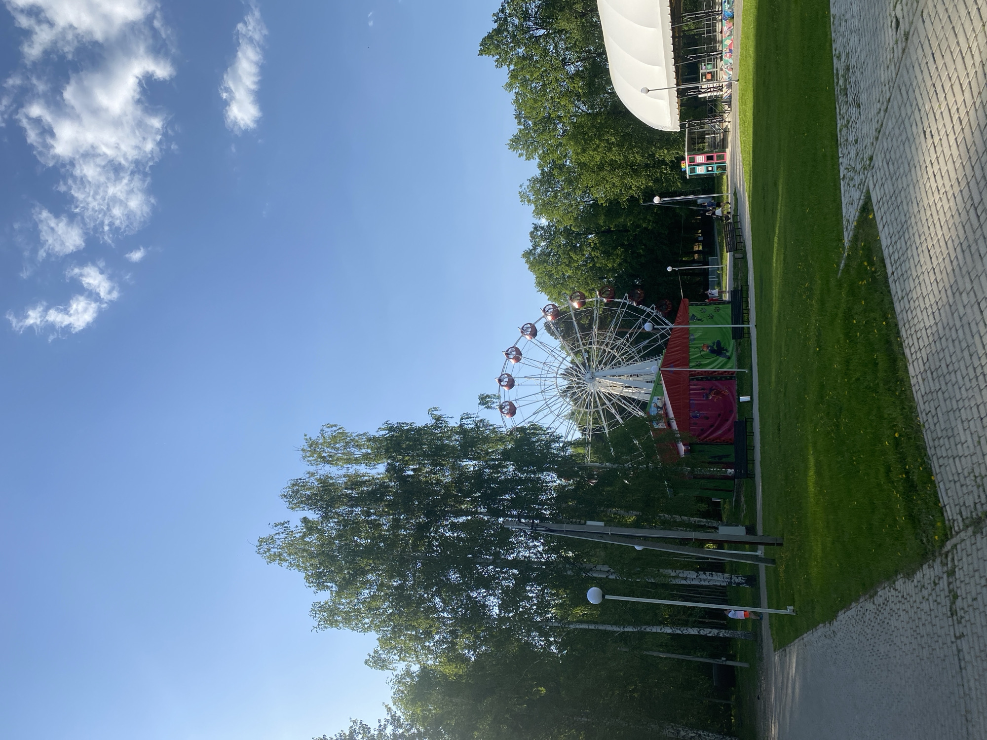 The image shows a Ferris wheel in a park setting on a sunny day with blue sky and scattered clouds. The location is likely Izhevsk, Russia. Tall green trees partially obscure the Ferris wheel. A white tent-like structure is visible in the background. There's a paved path on the right side of the image, next to a grassy area. Various flags and banners are present near the Ferris wheel. Several light posts are visible among the trees in front of the wheel. No people are clearly visible.
