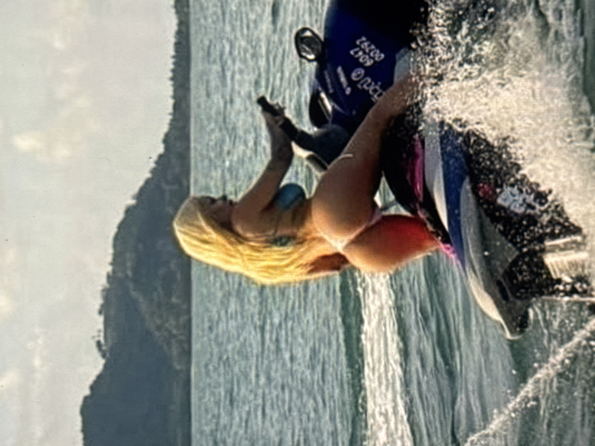 The media file depicts a woman with long blonde hair riding a jet ski in the ocean. She is wearing a bikini and is positioned on the back of the jet ski, appearing to hold onto a handle. The jet ski is in motion, creating a spray of water around it. In the background, a lush green hillside is visible, suggesting a tropical or coastal environment. The water is a clear blue, and there are ripples indicating movement. The lighting suggests it is daytime, possibly late afternoon given the warm light. The overall impression is one of recreation and enjoyment of a watersport. On the back of the jet ski, some text is visible in white on a dark blue background. It reads "26200 0047 2709" and also contains some circular logos.