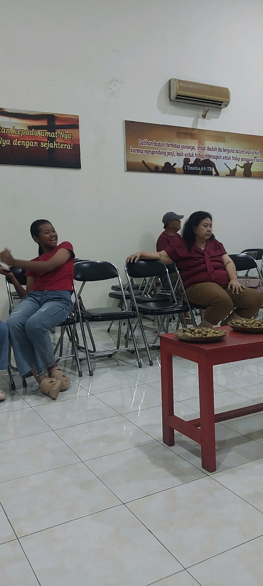 In Keling, Indonesia, an indoor scene depicts a gathering with several individuals seated on chairs. On the left, a young woman wearing a red shirt and ripped jeans is seated and appears to be laughing or speaking animatedly, with her arms raised. To her right, a man in a maroon shirt and a grey cap is seated, looking away from the camera. Further to the right, a woman in a maroon shirt and brown pants is seated, looking downwards with her hand resting on her lap. In the background, an air conditioning unit is mounted on the wall. Two framed posters are also visible on the wall, one on the left with text and a cross graphic, and another on the right with text and silhouettes of people. In the foreground, a red table holds two shallow bowls filled with what appear to be nuts or snacks. The floor is tiled with a light-colored pattern.