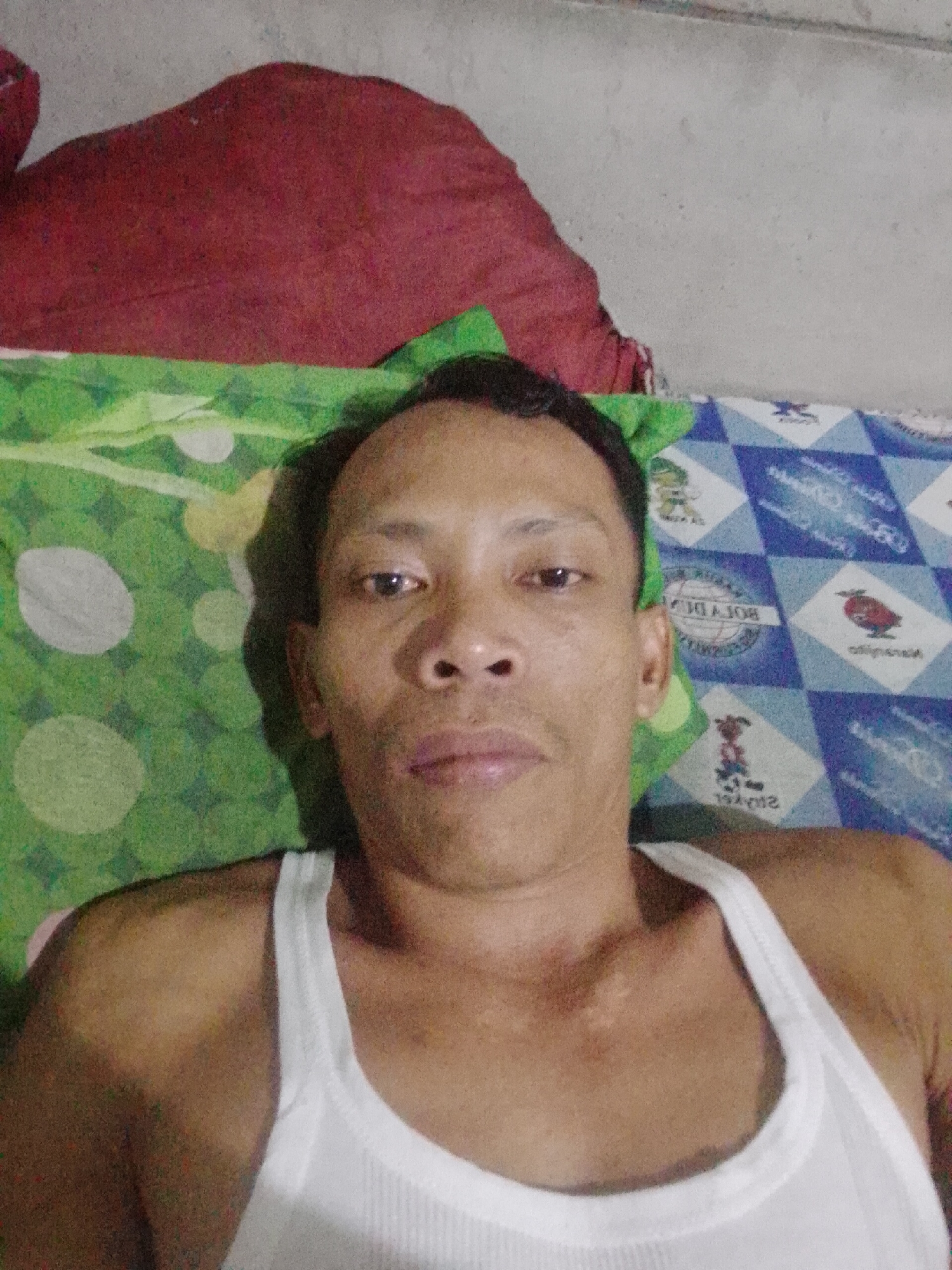 A man with short dark hair, light brown skin, and wearing a white ribbed tank top is lying on a bed. He is facing the camera. The bed has a green patterned pillow to his left, a blue patterned sheet on his right and a red pillow or blanket above his head. The location is likely Bukit Ketok, Indonesia, based on the prompt's context.