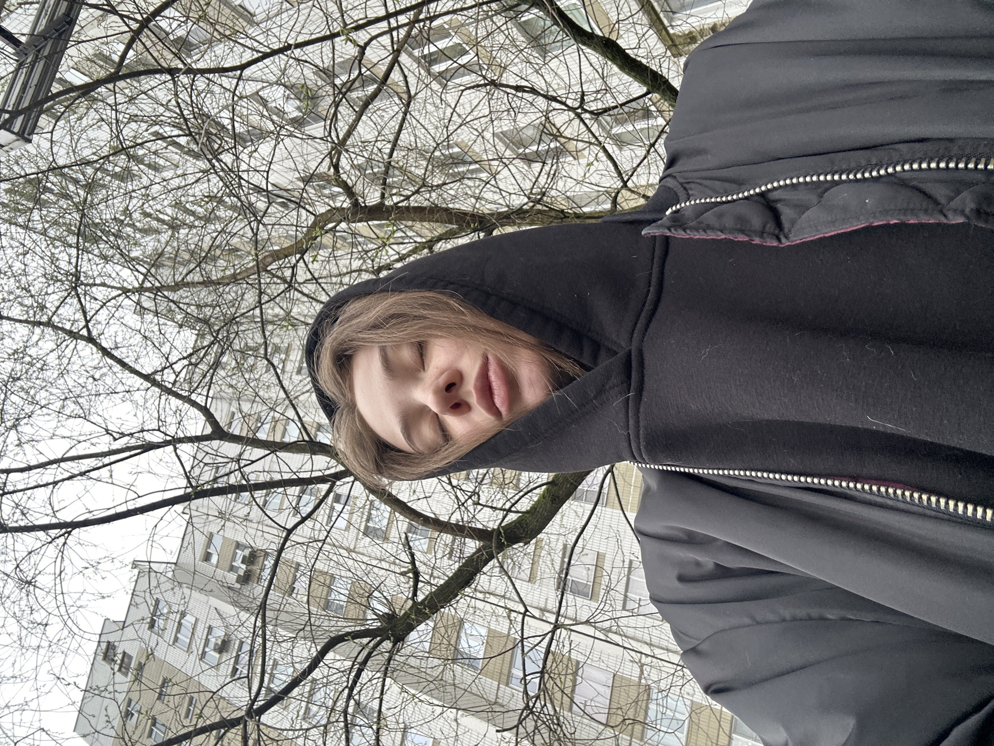 A person in a black hooded jacket looks down at the camera. Bare tree branches crisscross the frame against a pale sky. In the background is a multi-story building with repetitive window patterns, indicative of residential construction. The architecture and appearance of the building align with typical structures found in Moscow, Russia.