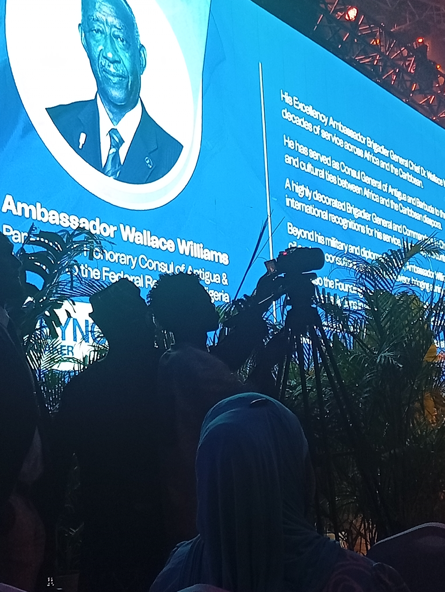A photograph shows a man on a screen identified as Ambassador Wallace Williams. The photograph appears to be taken at an event honoring him, judging by the description of his accomplishments and titles on the screen. Several people are in the foreground watching the screen, including one wearing a hijab. There is a video camera set up in front of the screen and palm plants are also in the foreground, partially obscuring the camera and some of the people.