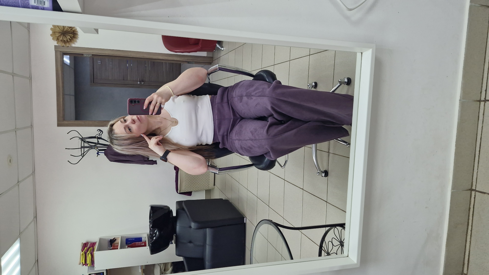 A mirror reflects a scene inside what appears to be a hair salon. A woman is sitting in a salon chair, taking a selfie with a smartphone. She wears a white top and purple pants. Visible elements in the salon include a hair washing station, a coat rack, hair products on a shelf, and a red chair. The floor is tiled. The location is potentially in Otchor, Russia.