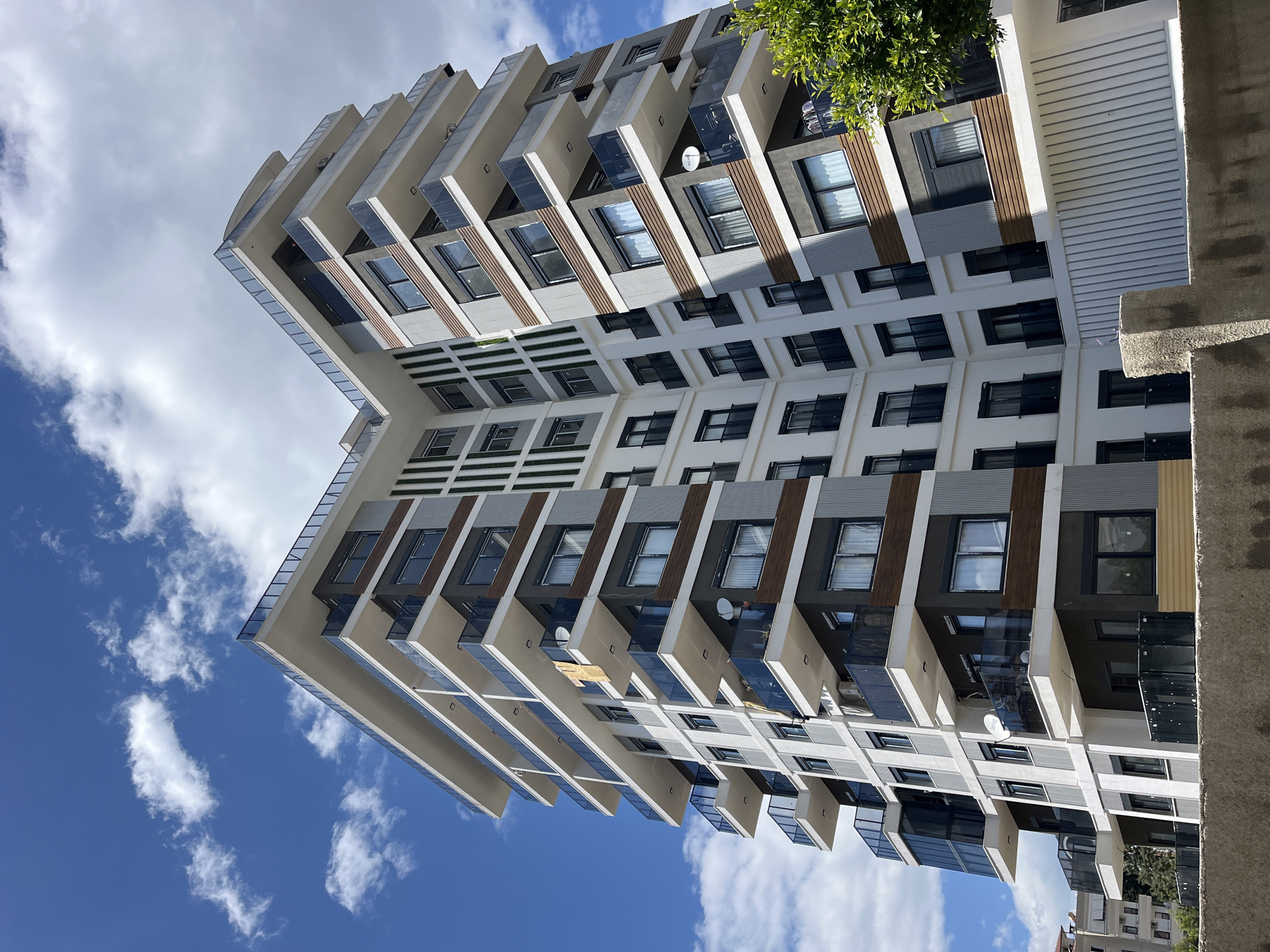 The image shows a multi-story building in Alanya, Turkey. The structure features a modern architectural style with projecting balconies and glass panels. The building's facade incorporates a combination of white, grey, and wood-toned elements. The sky above is blue with scattered white clouds. The image is taken from a low angle, looking upward at the building.