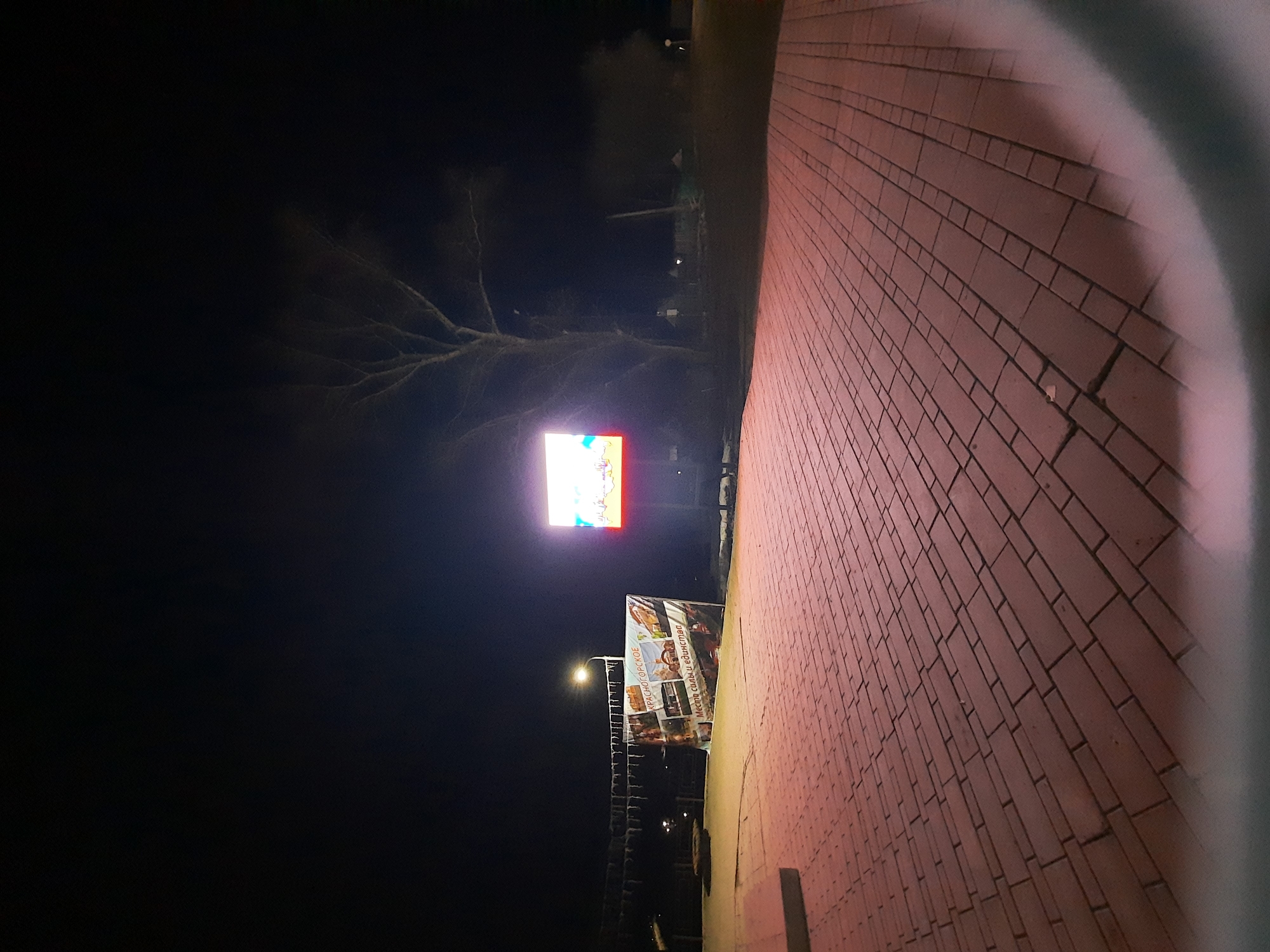 The image shows an outdoor scene at night in Krasnogorskoye, Russia. A brick walkway is visible in the foreground, leading towards an illuminated sign displaying text in Russian: "КРАСНОГОРСКОЕ - МЕСТО СИЛЫ И ЕДИНСТВА". Above the sign, there is an electronic billboard with a brightly lit display. A street lamp is also illuminated nearby. The background is dark, with trees and other structures faintly visible. The overall impression is of a public area at night with illuminated signage.