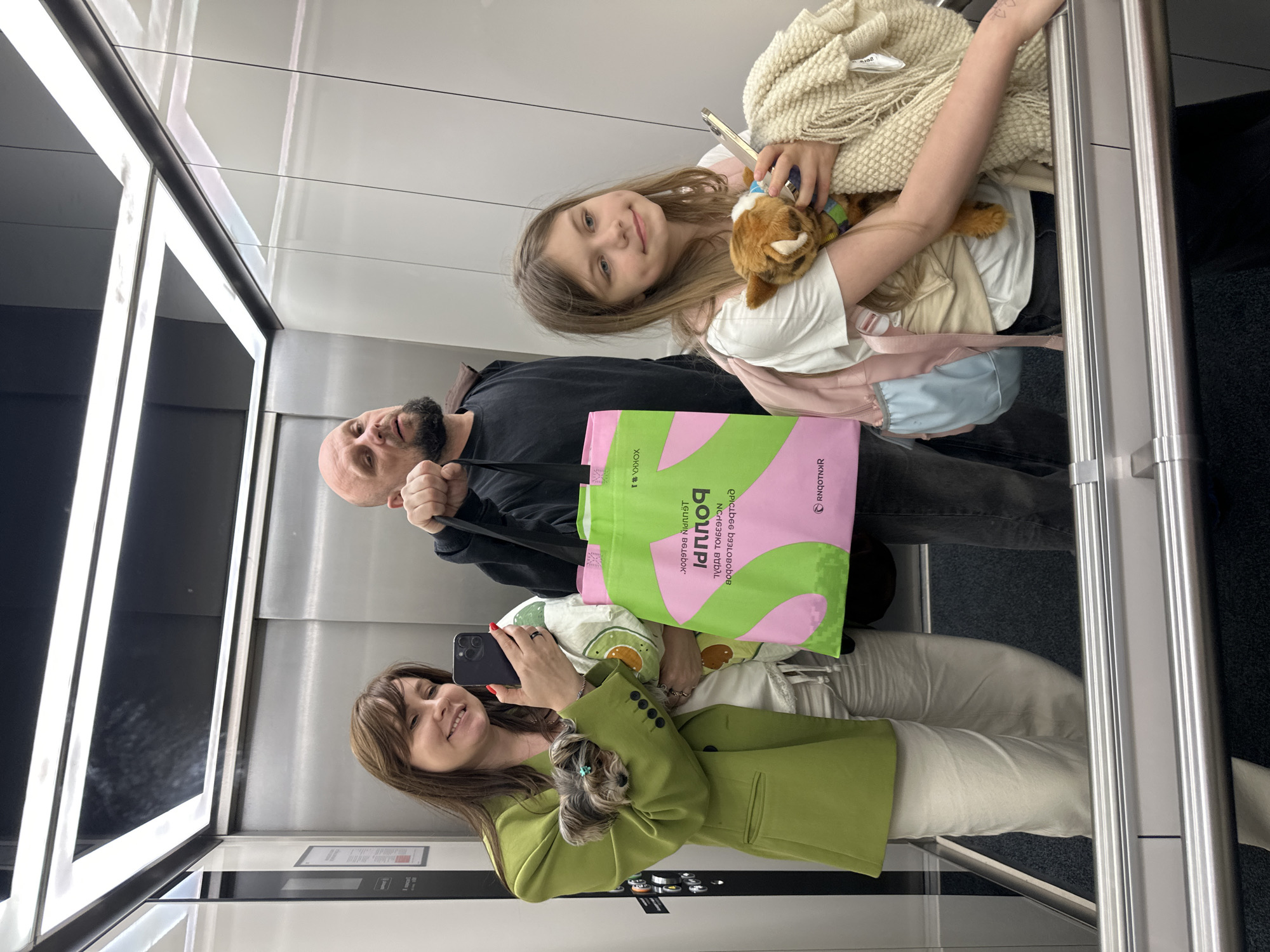 Captured in an elevator, this image depicts three individuals: a young girl holding a plush toy and wrapped in a blanket, a man with a shaved head carrying a shopping bag, and a woman in a green jacket holding a small dog while taking a picture with her smartphone. The shopping bag displays Cyrillic text, indicating the location is likely in Moscow, Russia. The elevator has modern steel panels and artificial lighting.