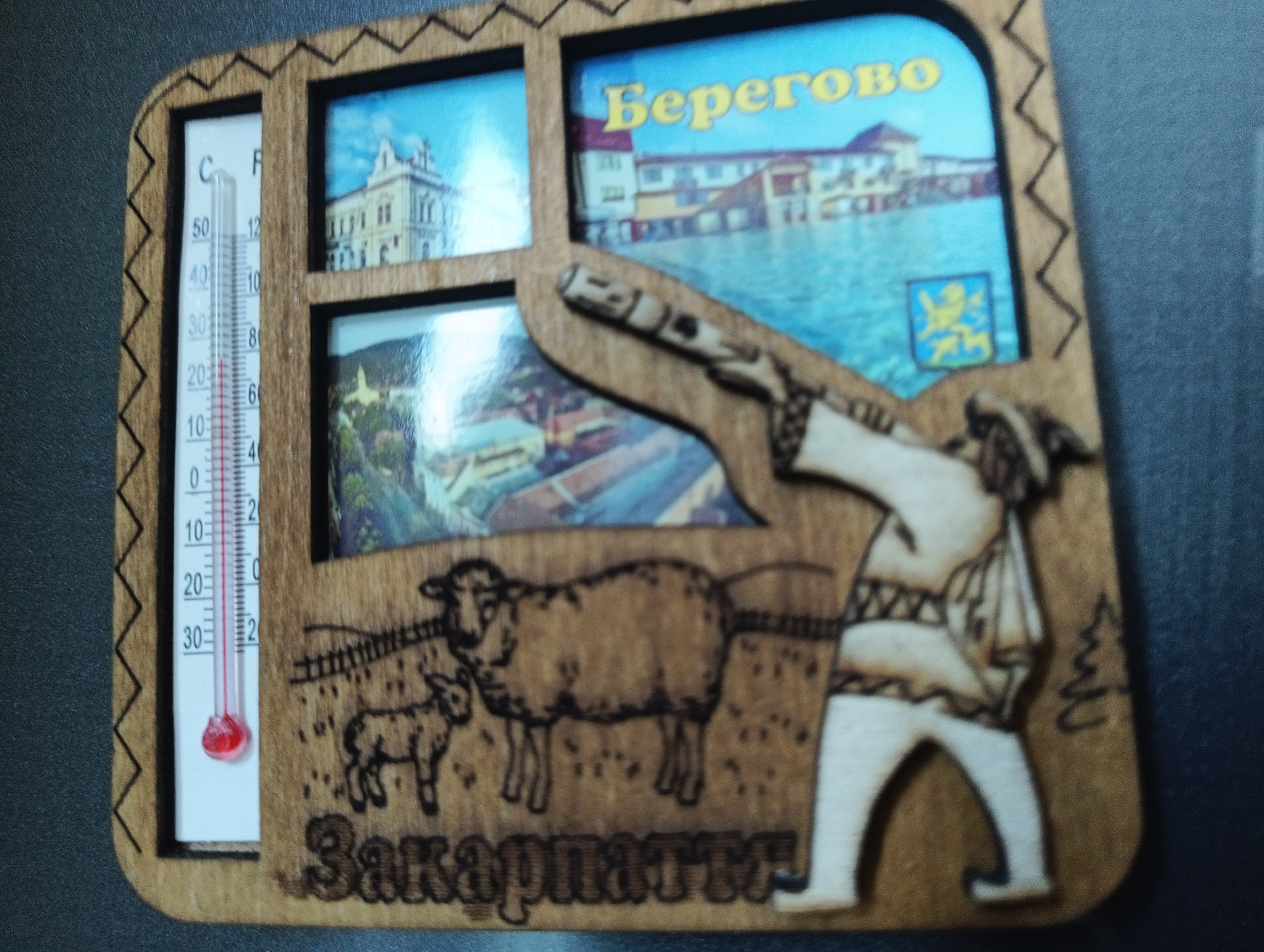 The image displays a wooden souvenir magnet. The magnet is shaped like a house with a zig-zag patterned frame. It features a thermometer on the left, showing a temperature of approximately 15 degrees Celsius. To the right of the thermometer are two small framed pictures. The top picture depicts a building with classical architecture, and the bottom picture shows a village scene with trees and buildings.
Overlapping the framed pictures and extending to the right is a larger, un-framed image also printed on the magnet. This image shows a body of water with buildings along its edge, and text that appears to read "Beregovo" in yellow Cyrillic letters. Below this, there is a coat of arms with a lion.
Further down, on the wooden part of the magnet, there are carved images. In the center, there are two sheep, one adult and one lamb. To the right of the sheep is a carved figure of a person in traditional clothing, holding a wooden instrument. Below the sheep and the carved figure, there is text in Cyrillic letters that appears to read "Zakarpatt".
The overall context is a tourist souvenir, likely from a location named Beregovo, possibly related to the Zakarpatt region. There is no information in the image that places this souvenir in the city of Karnobat, Bulgaria.